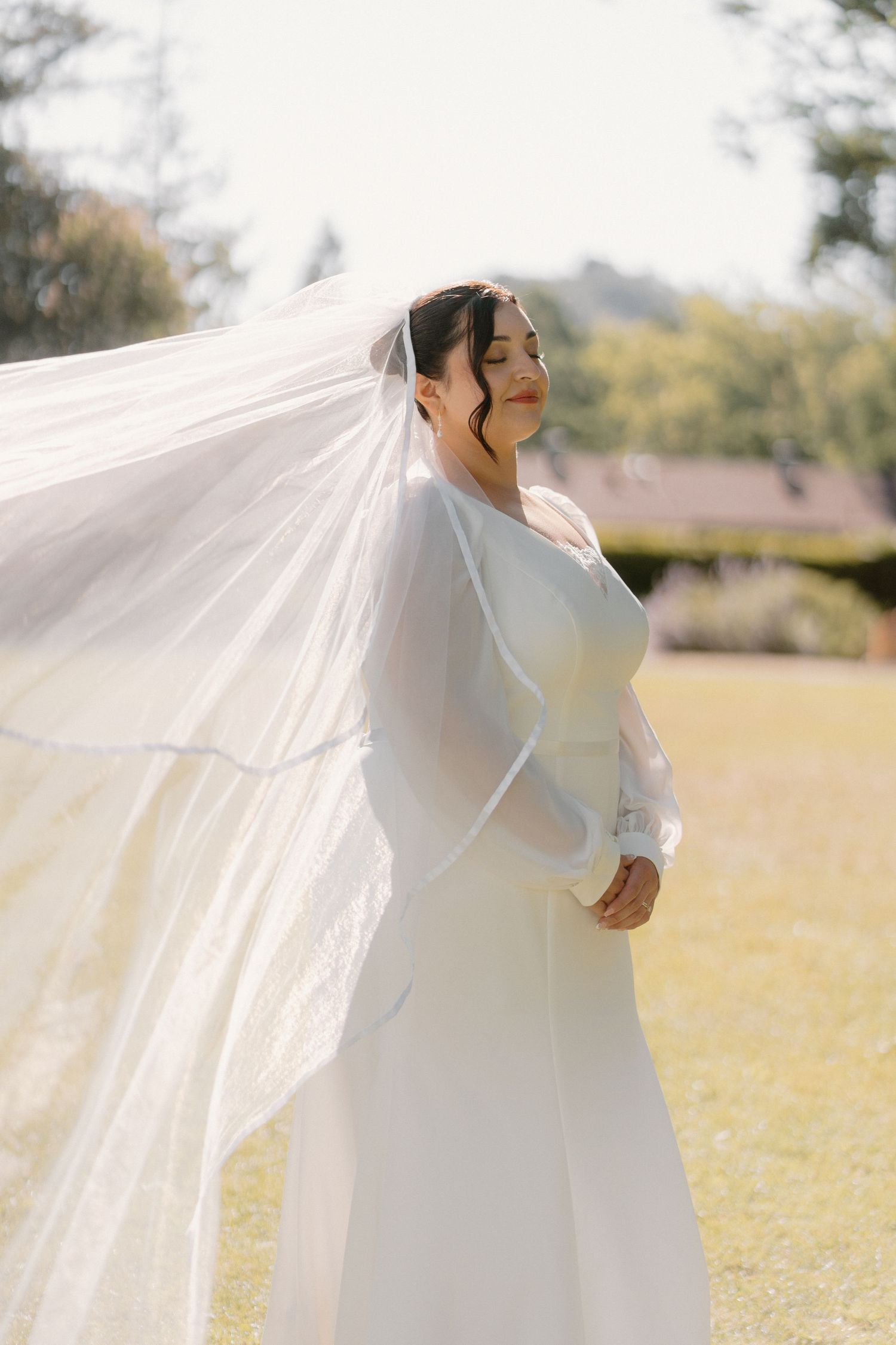 Bride in white long-sleeve gown with flowing veil billowing in breeze on sunny lawn.