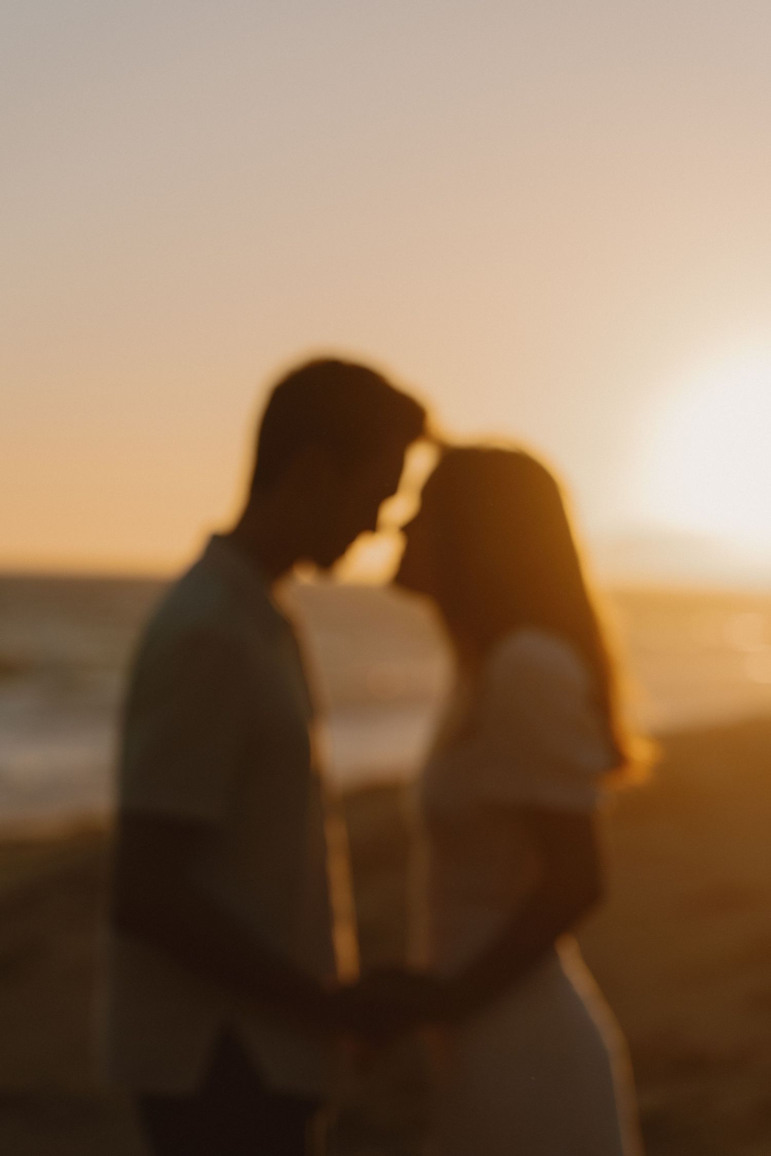Blurry silhouette of a romantic couple almost kissing at golden hour on a beach at sunset.