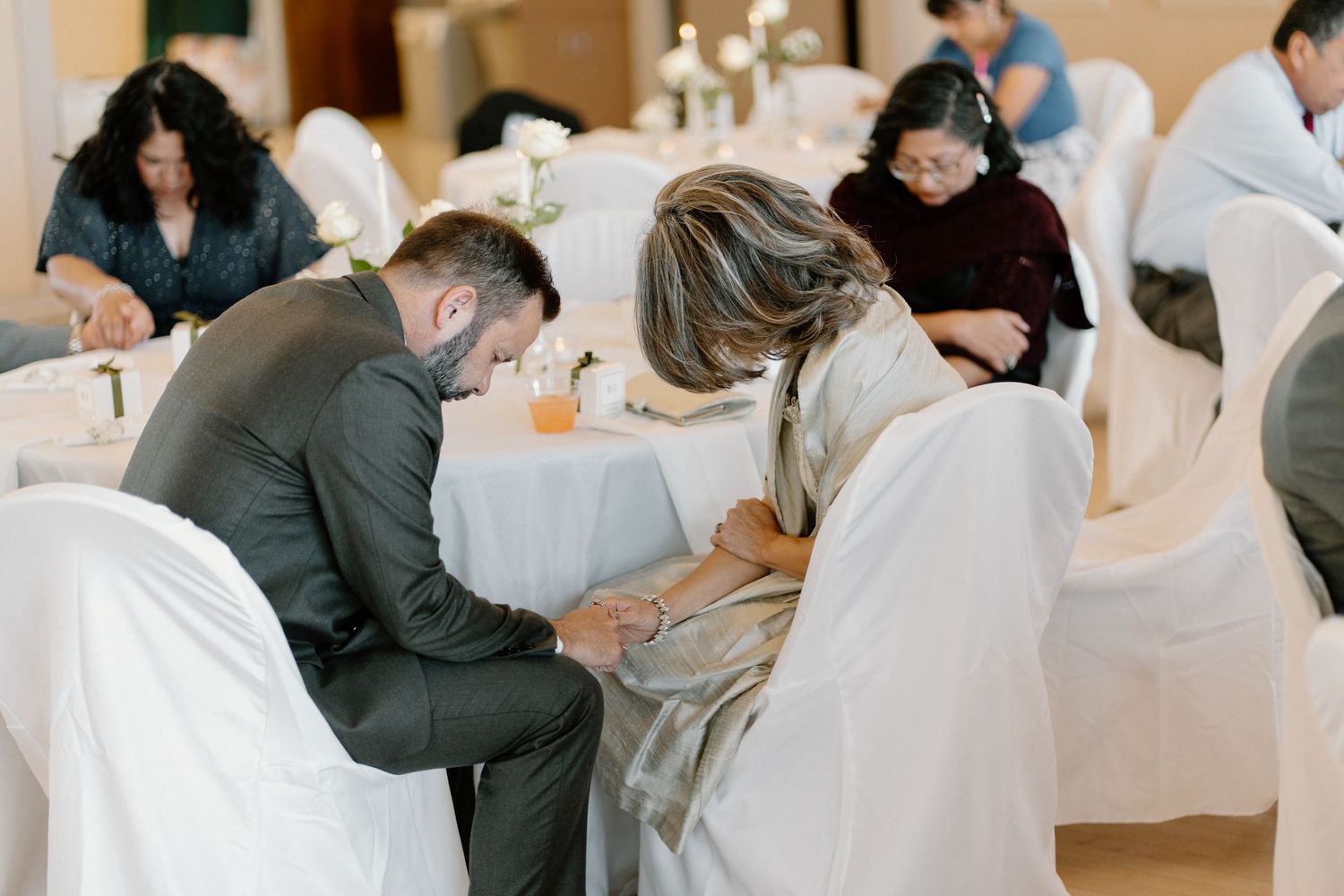 Wedding guests sign items at white cloth-covered tables in elegant reception hall with floral centerpieces.