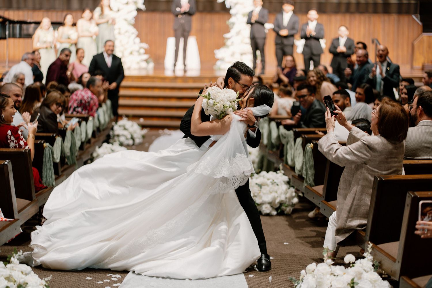 Groom dips bride in elegant white gown during romantic wedding ceremony recessional with floral decorations.