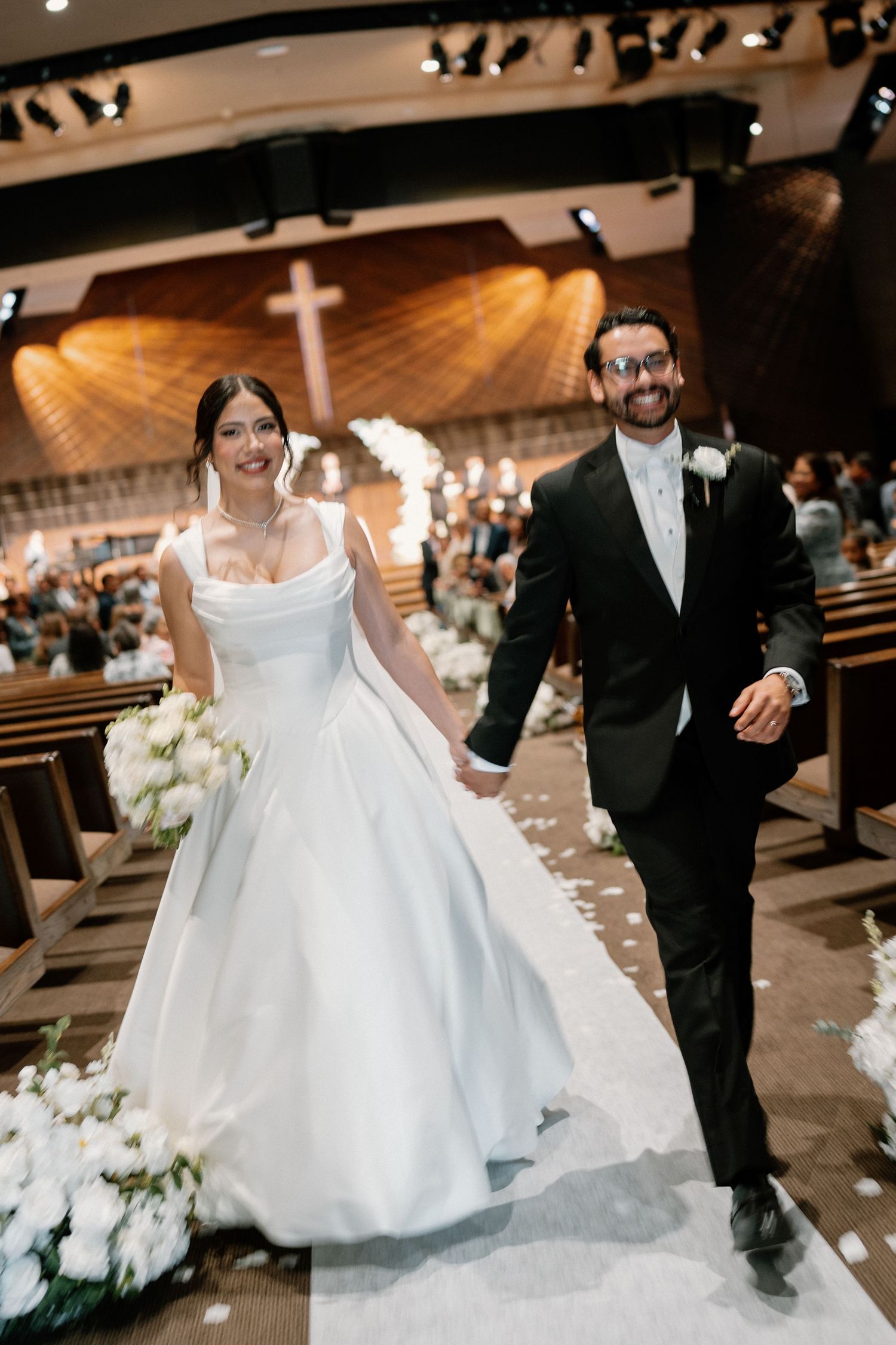 Newlyweds walk down the church aisle smiling, bride in white gown holding bouquet, groom in black tuxedo.