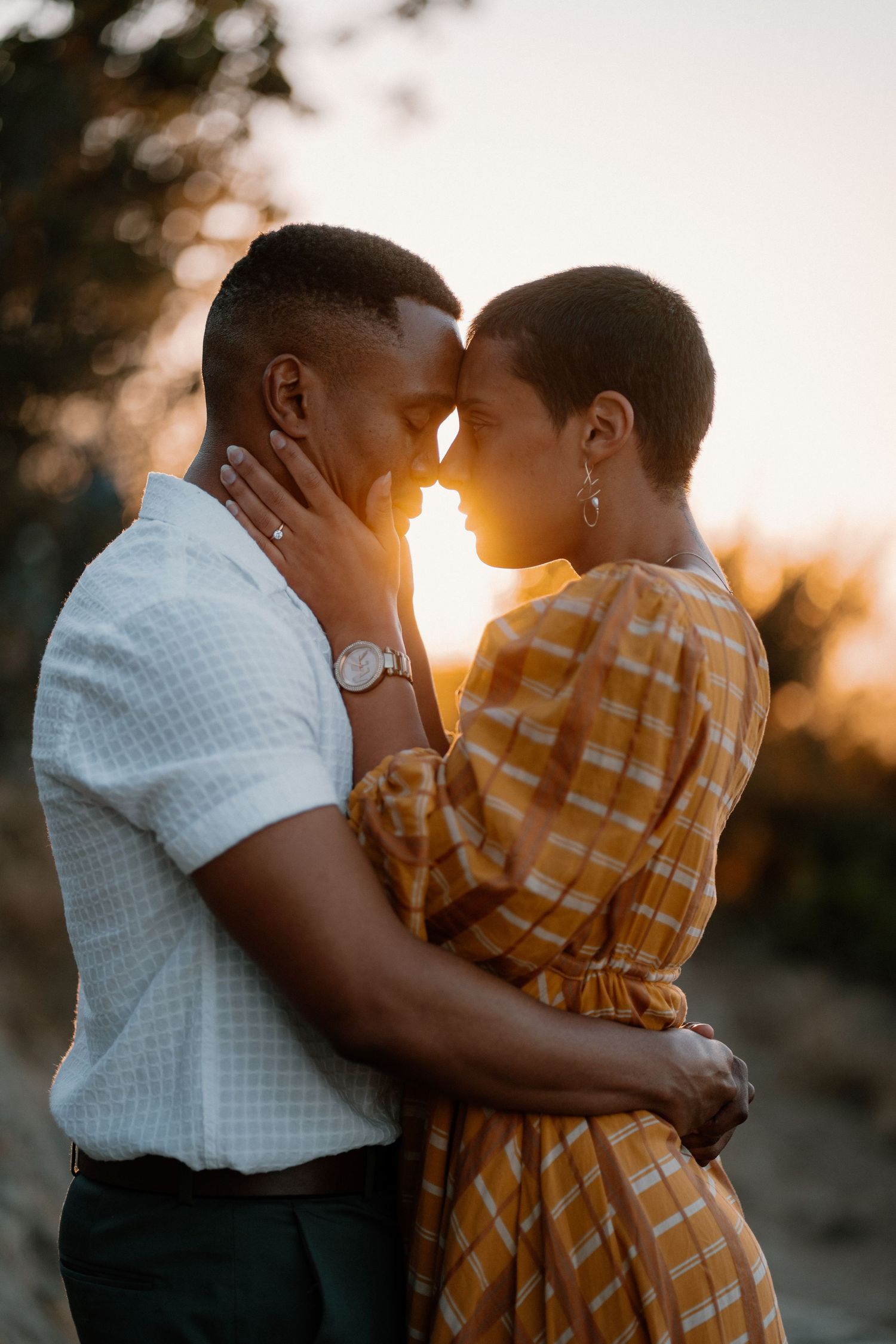 Romantic couple touching foreheads at sunset, woman in yellow plaid dress and man in white shirt embracing.