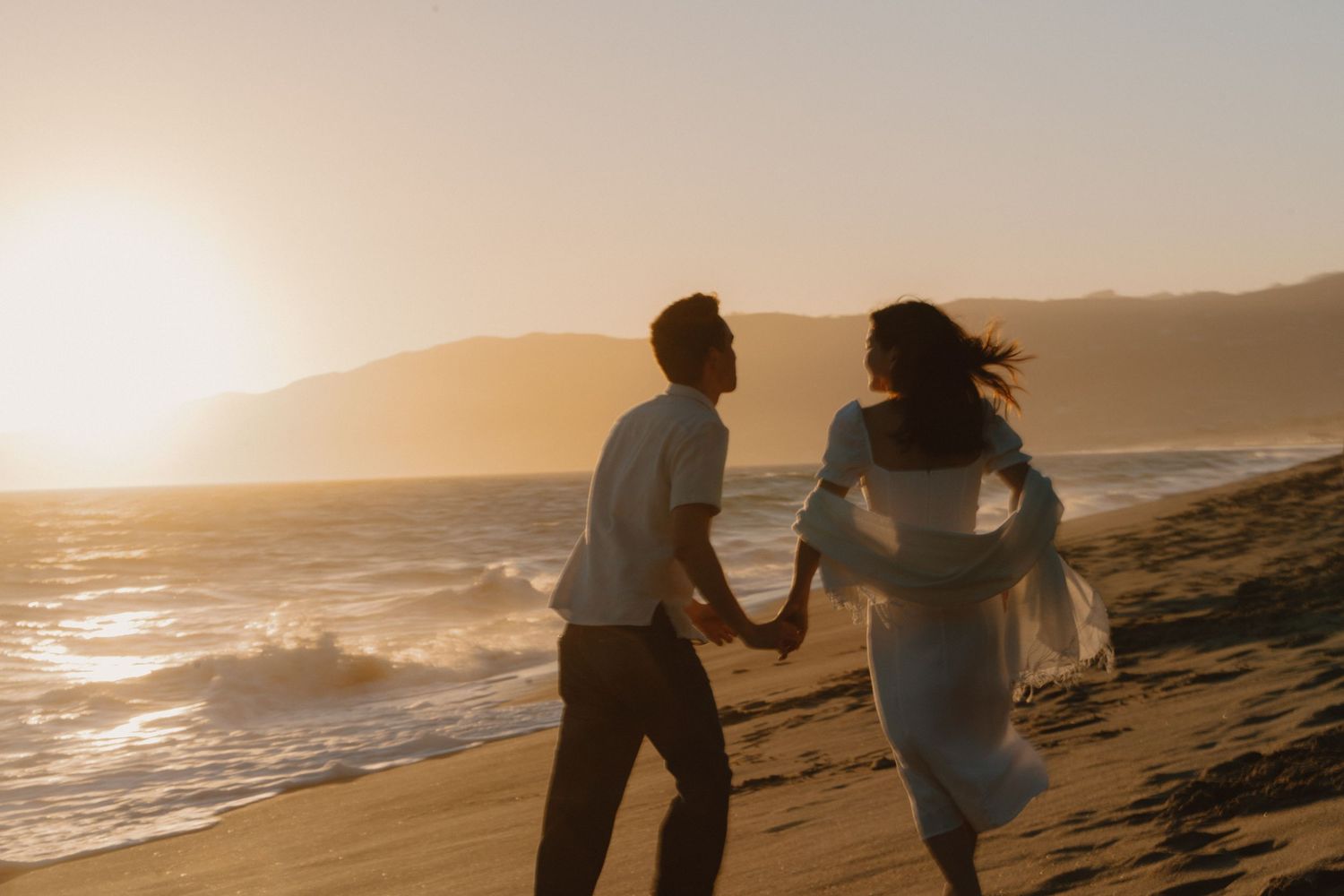 Couple holding hands running along sandy beach at golden sunset.