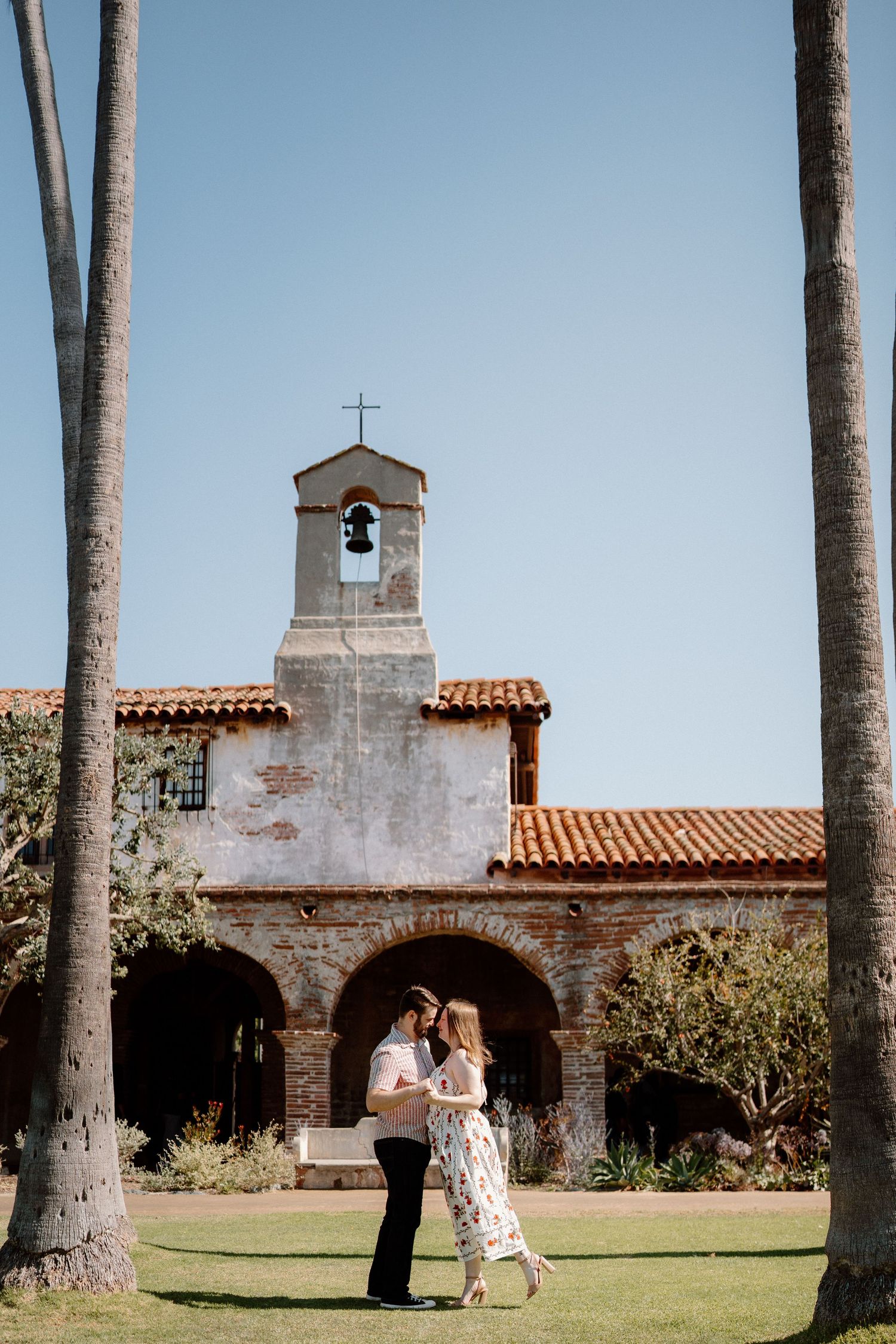 Couple dancing on lawn before a historic Spanish mission church with bell tower and palm trees.