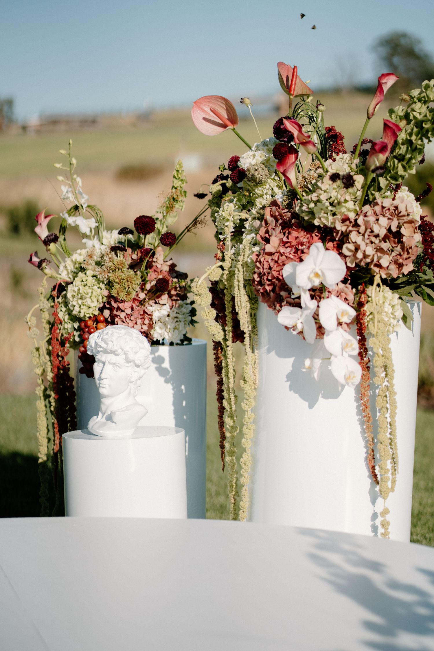 Elegant wedding floral arrangements with burgundy and mauve blooms beside a white classical bust.