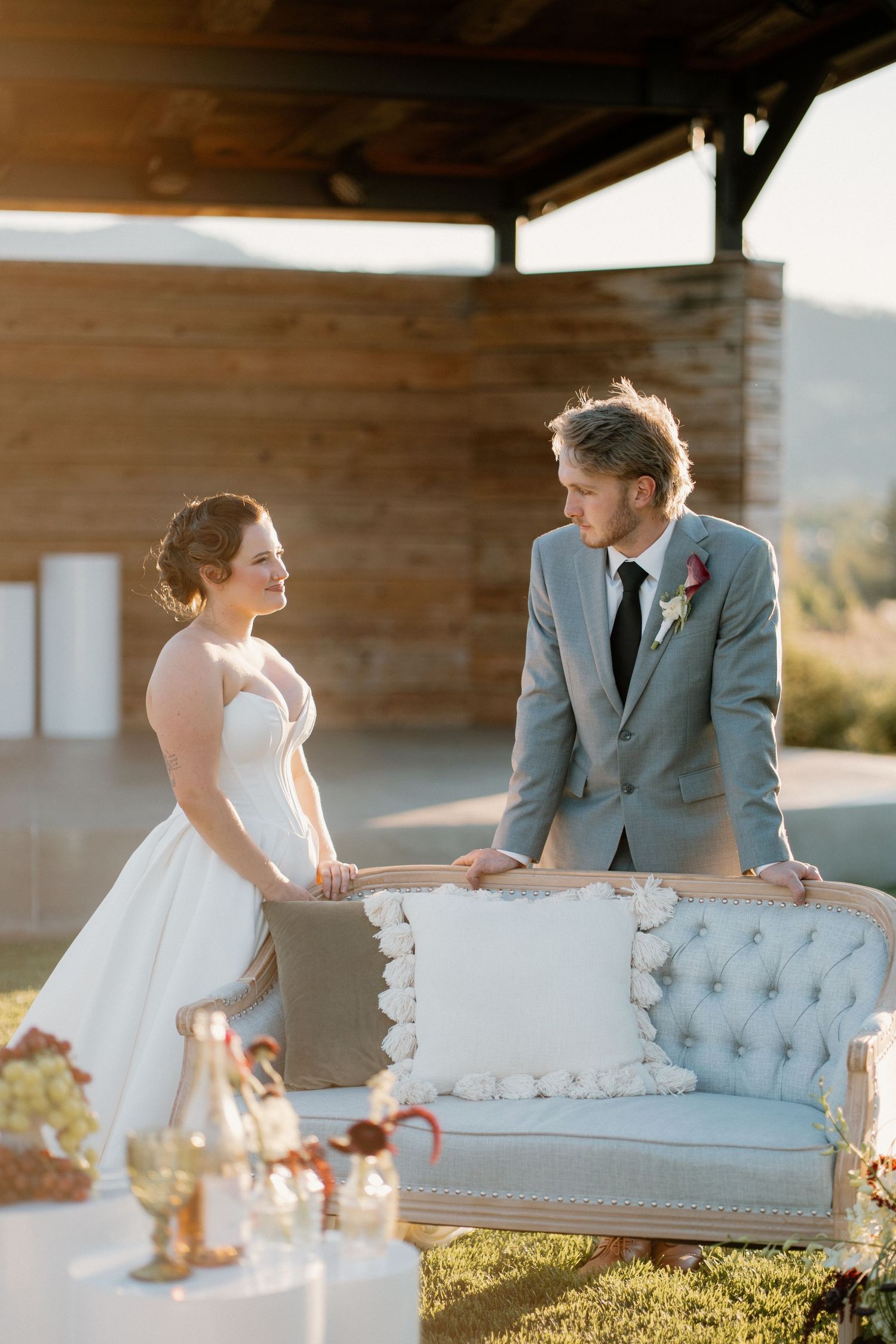 Bride and groom share a smile leaning over a tufted sofa at golden hour outdoor reception.