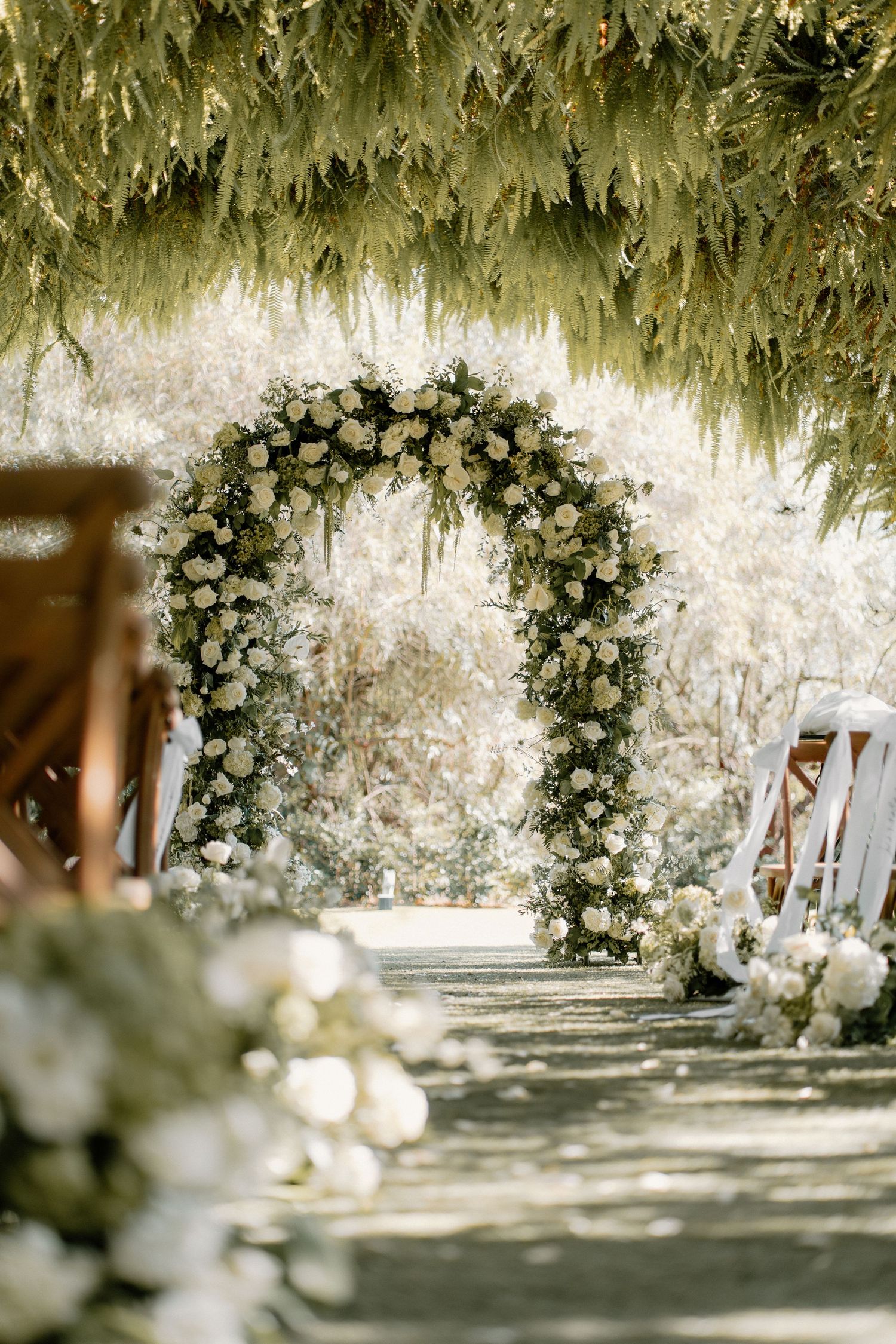 Stunning white floral arch framed by hanging greenery along a sunlit outdoor wedding aisle.