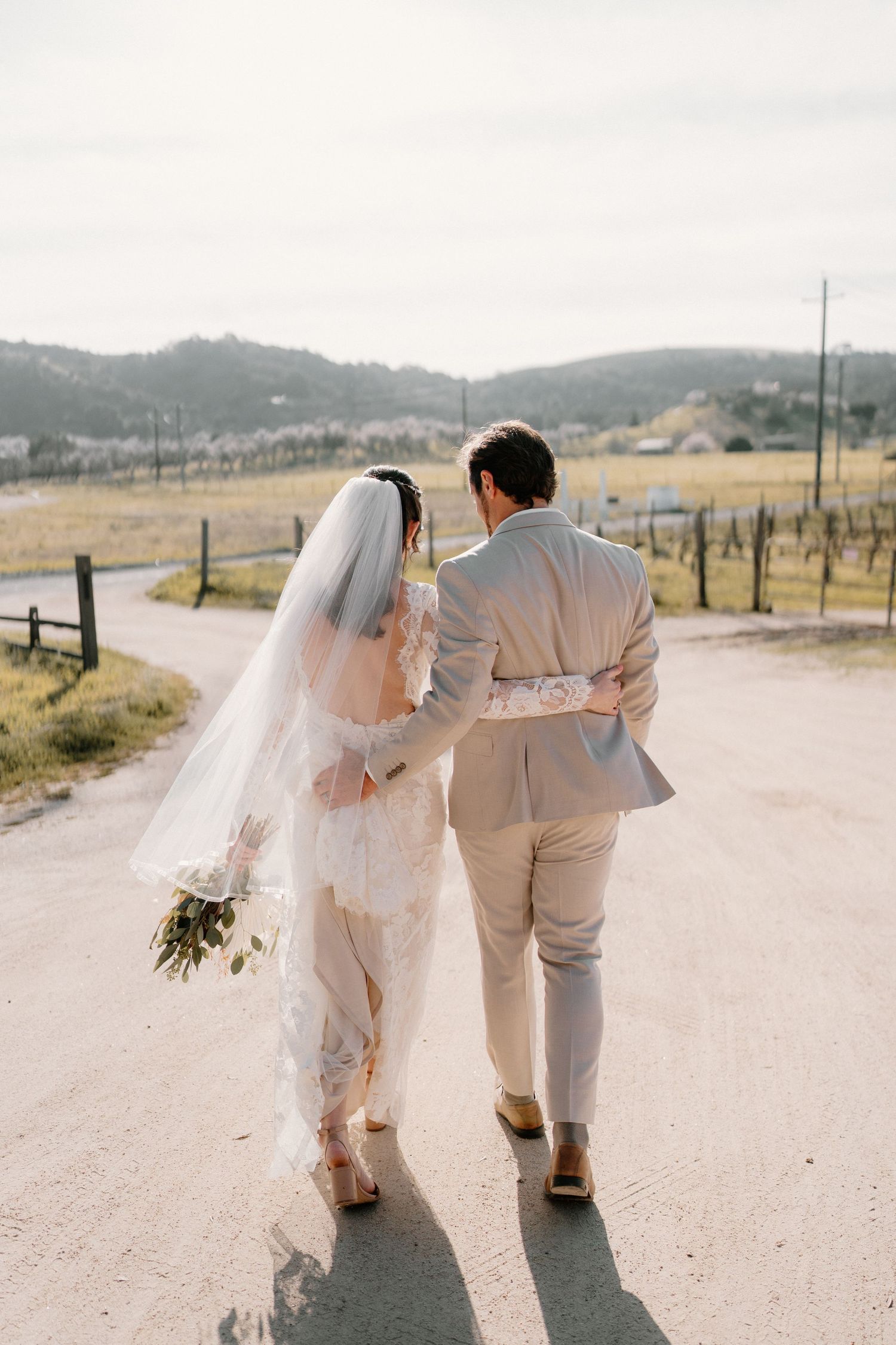 Bride and groom walking away hand-in-hand on dirt road with vineyard and rolling hills backdrop.