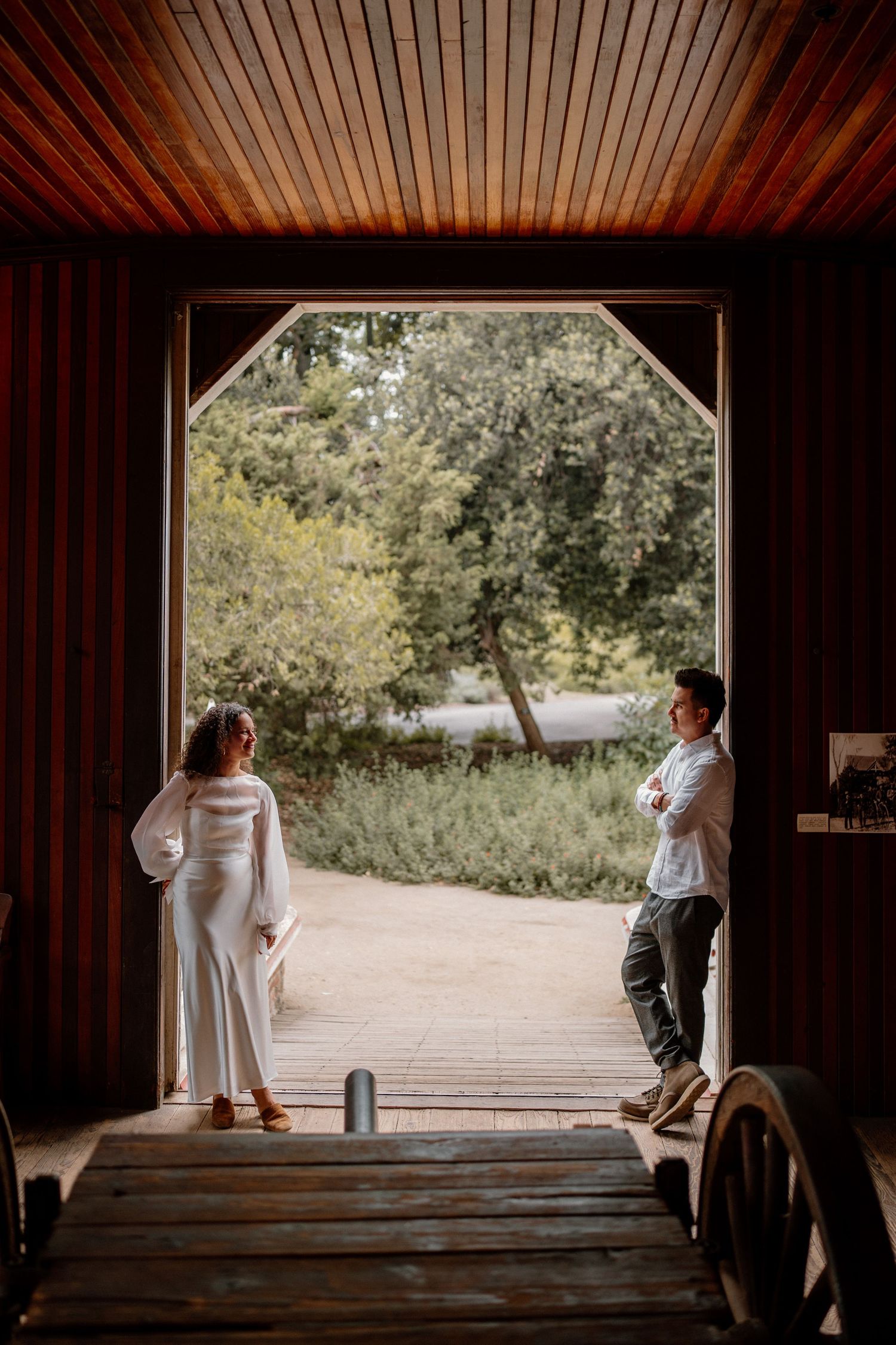 A bride and groom stand apart in a rustic barn doorway framed by lush greenery outside.