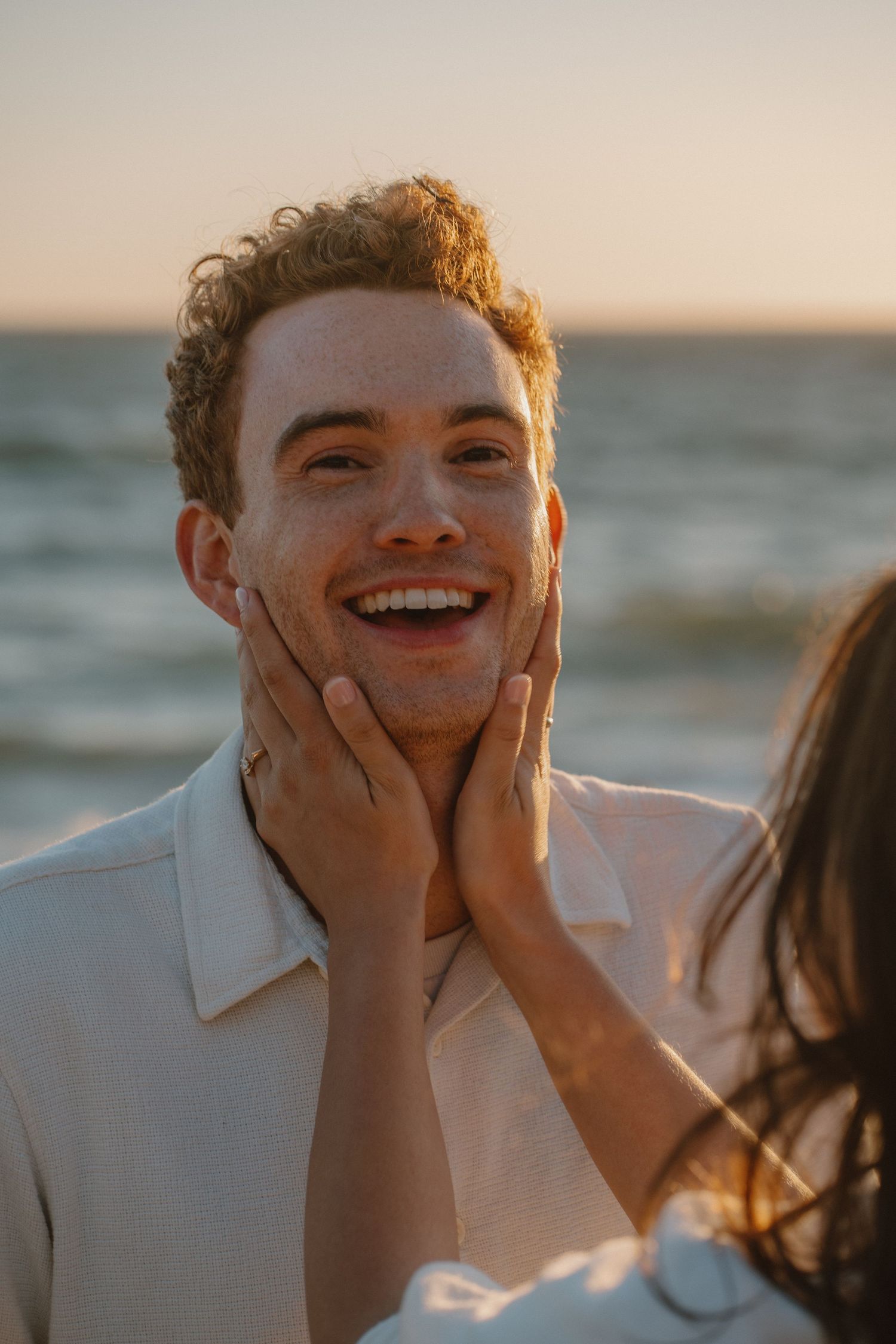 Smiling freckled man with curly hair being held by a woman's hands on his face at golden hour beach.