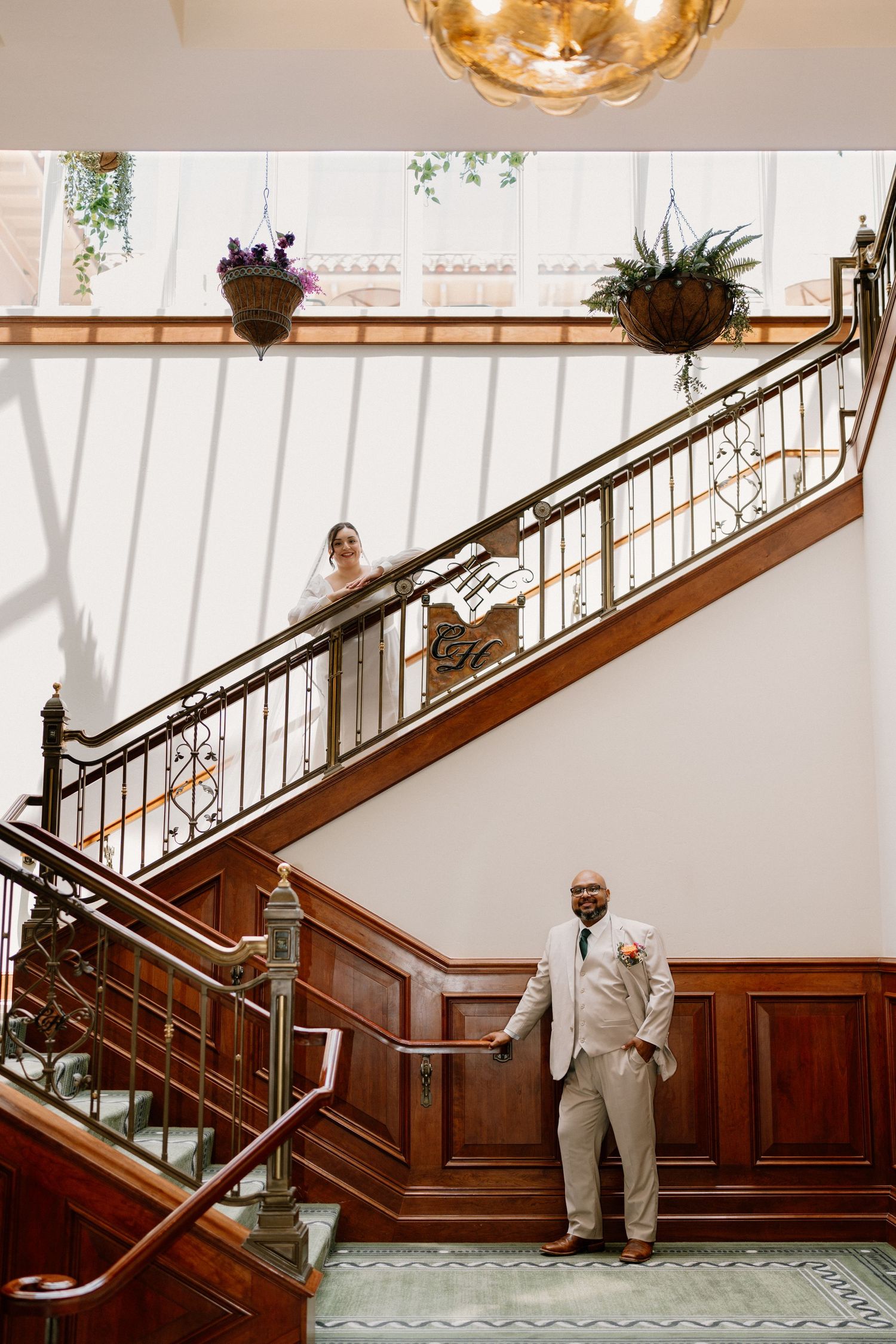 Wedding couple on grand wooden staircase, groom below in beige suit, bride above in white dress.