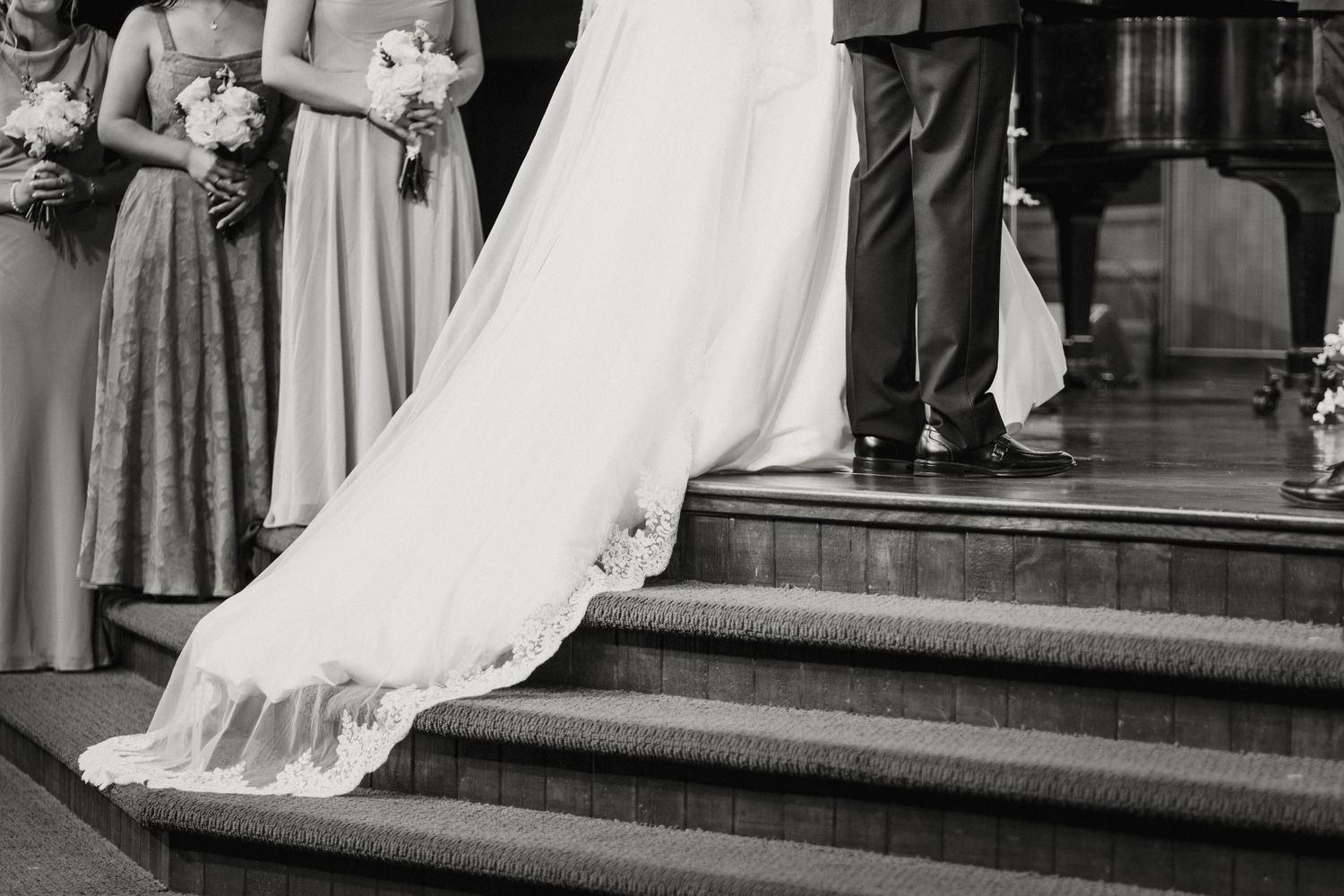 Black and white photo of a bride's flowing wedding gown train on church steps with bridesmaids.