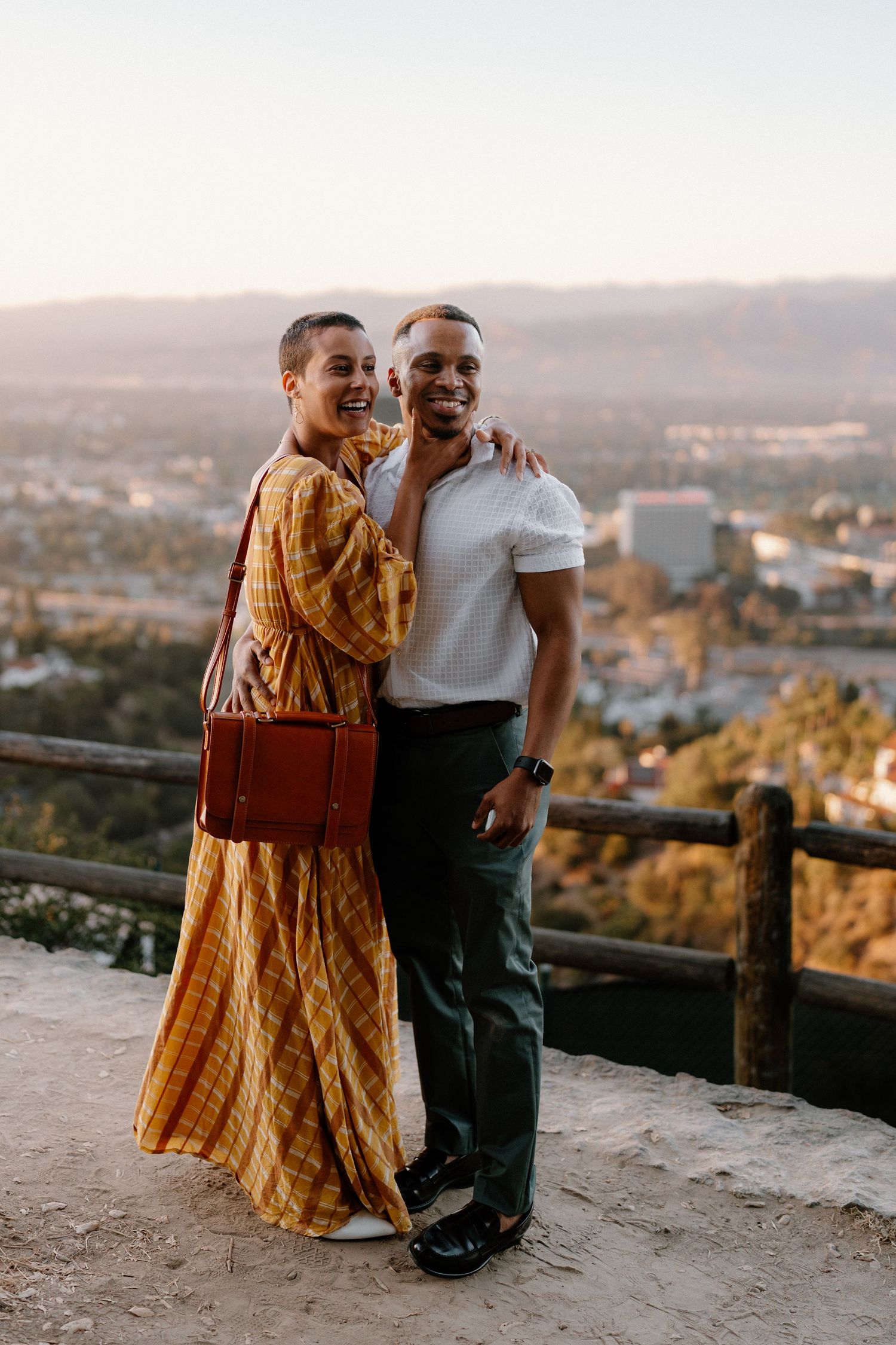 Couple posing together at a scenic overlook with city and mountain views at sunset, woman in yellow dress.