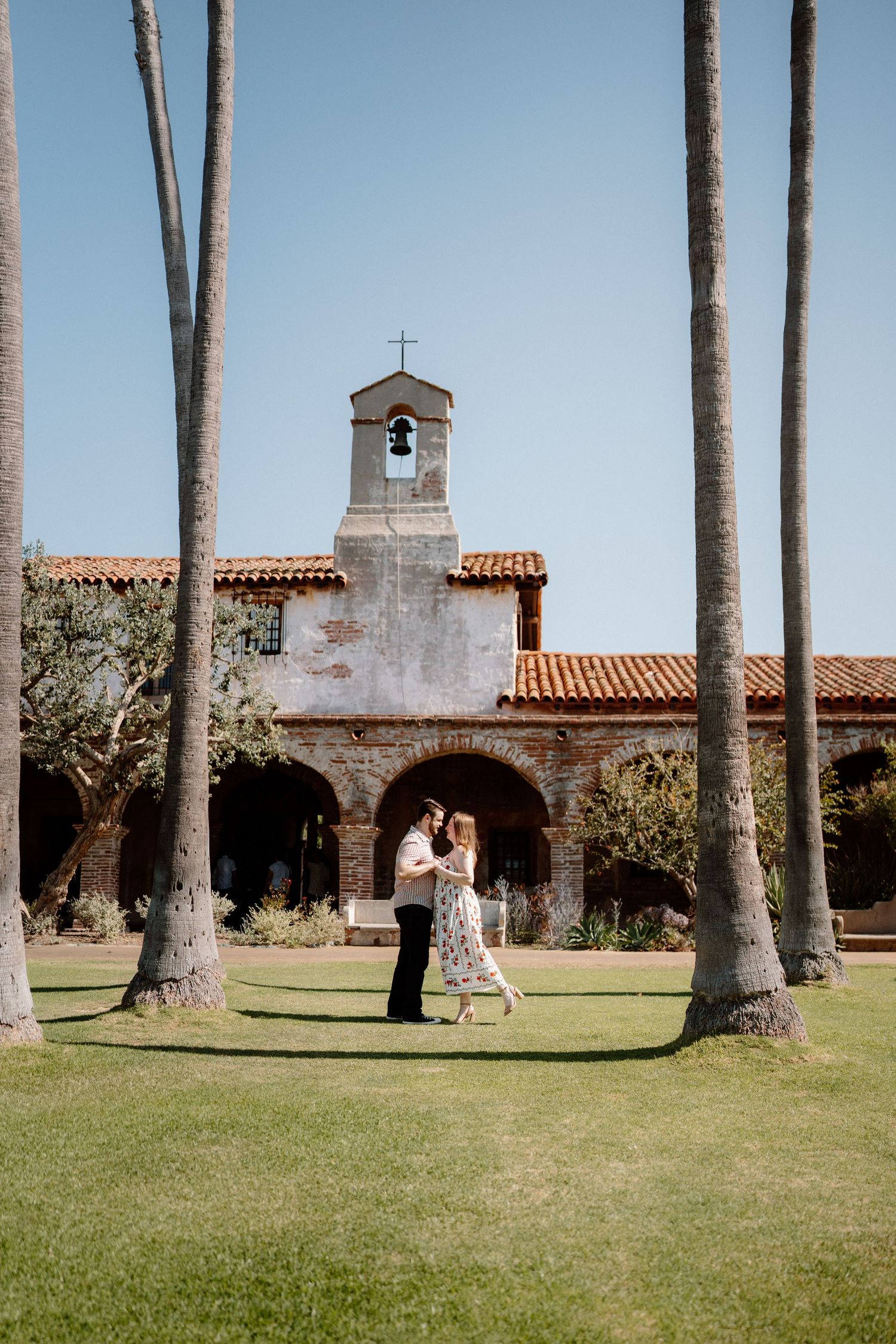 Couple dancing on green lawn before a historic Spanish mission bell tower framed by tall palm trees.