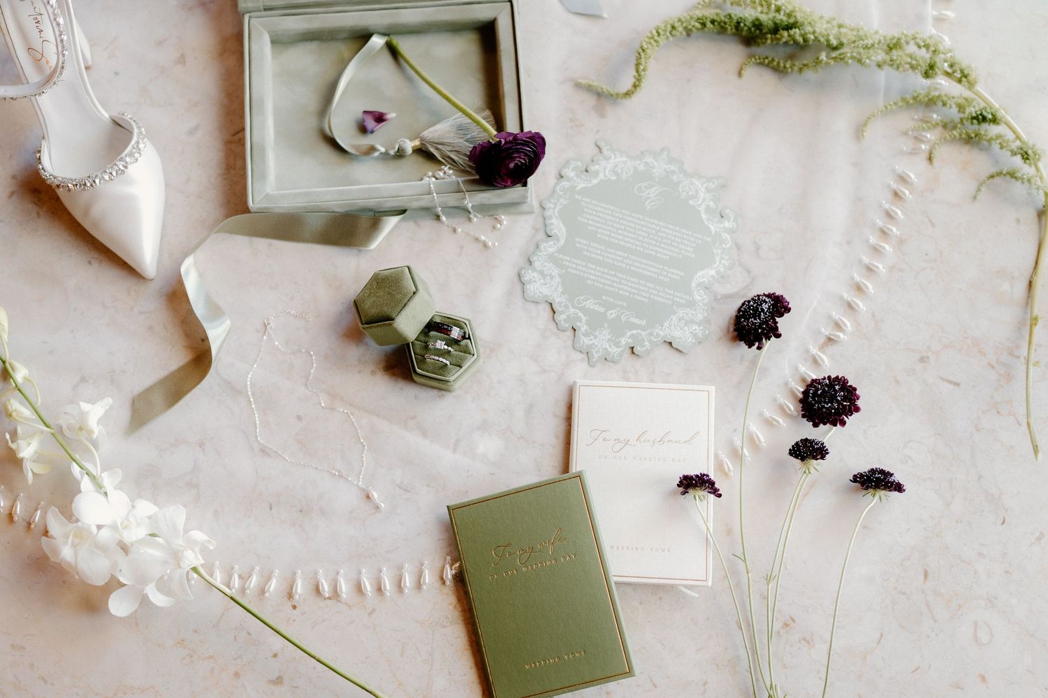 Elegant wedding flat lay with sage green stationery, ring box, dried flowers, and bridal accessories on marble.
