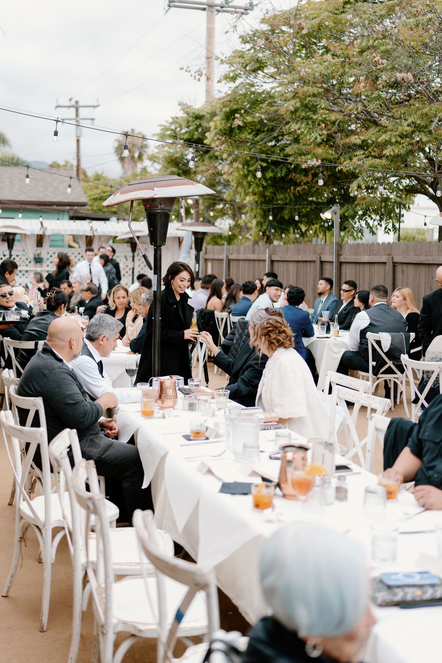 Guests seated at white-clothed tables during an outdoor wedding reception while a woman gives a speech.