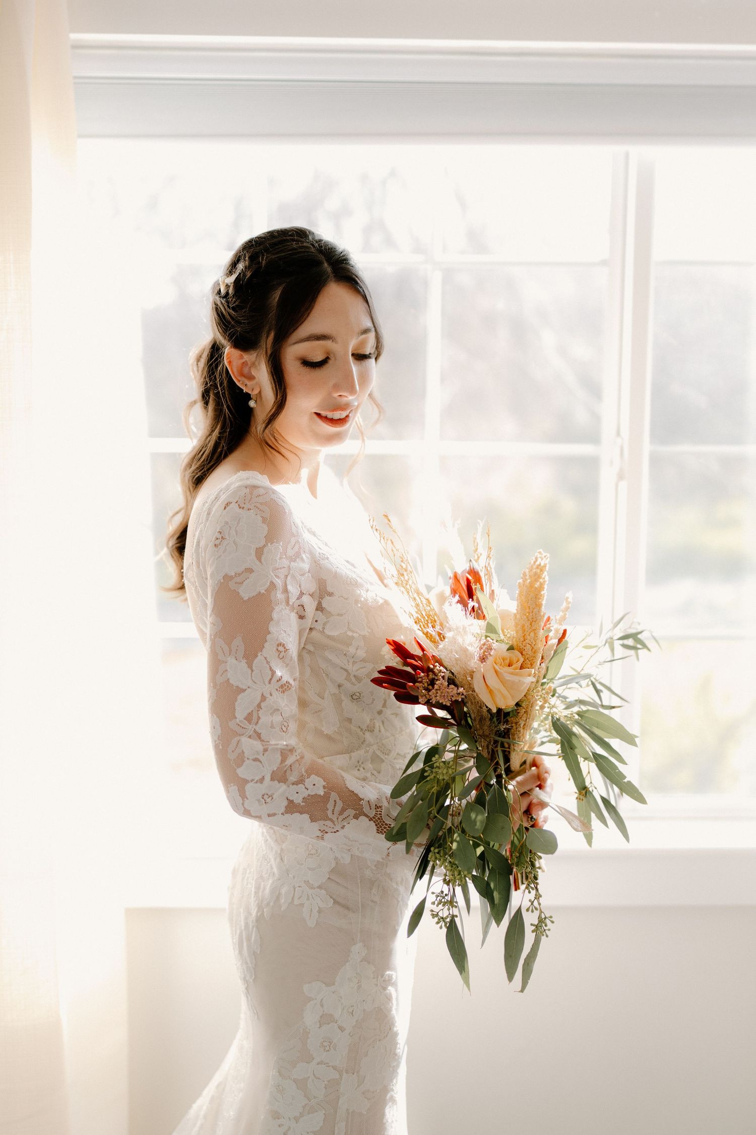 Bride in lace long-sleeve wedding gown holding boho bouquet with dried florals standing by sunlit window.