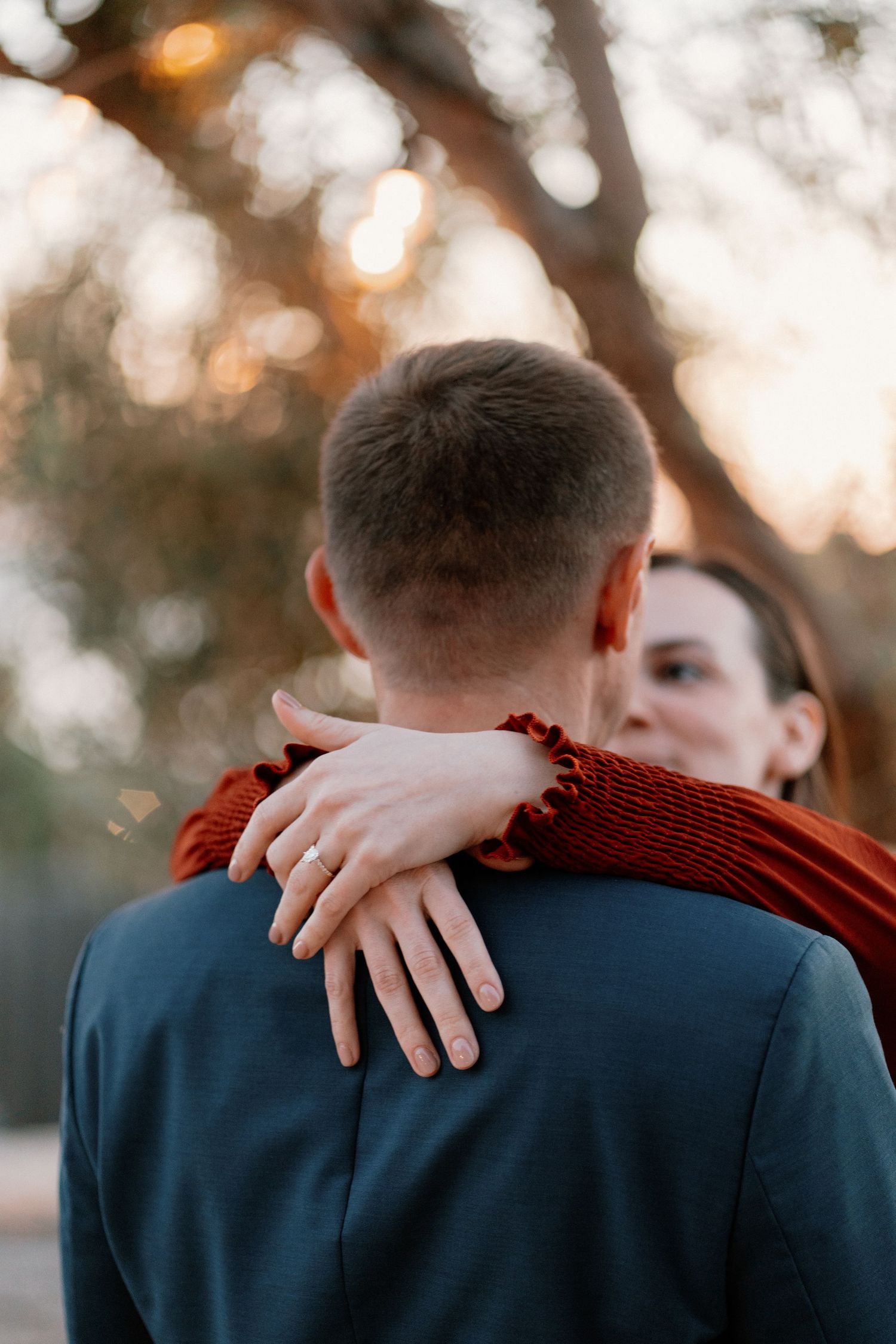 Couple embracing outdoors at sunset, woman in red dress showing engagement ring on man's shoulder.