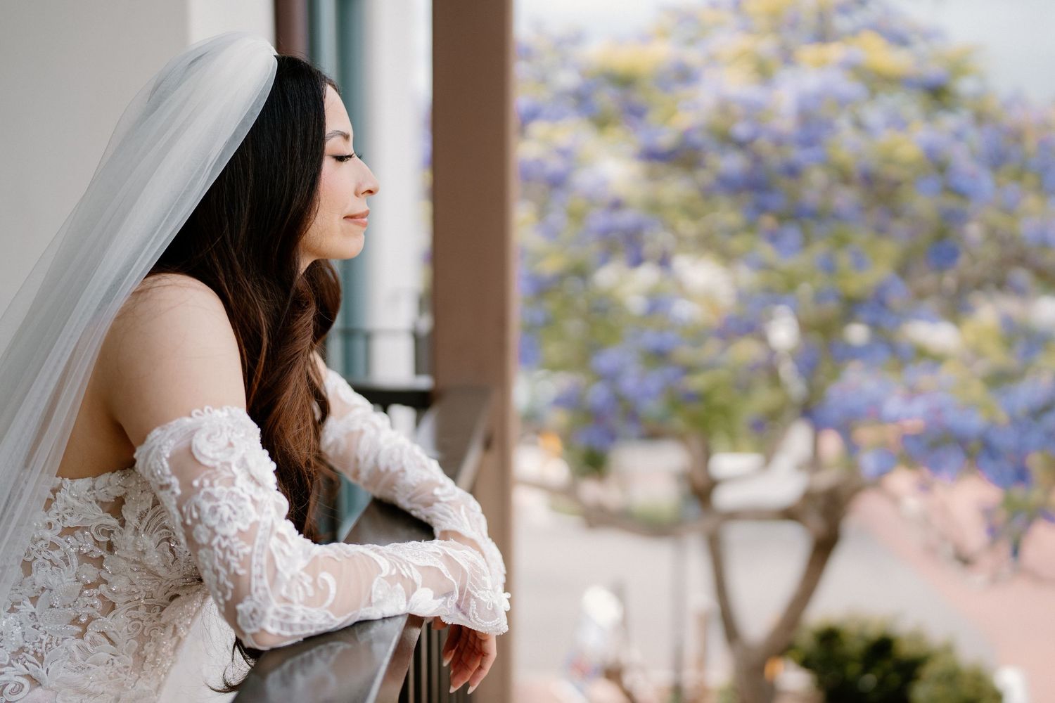Bride in lace off-shoulder gown and veil gazes pensively from a balcony with blooming purple trees.