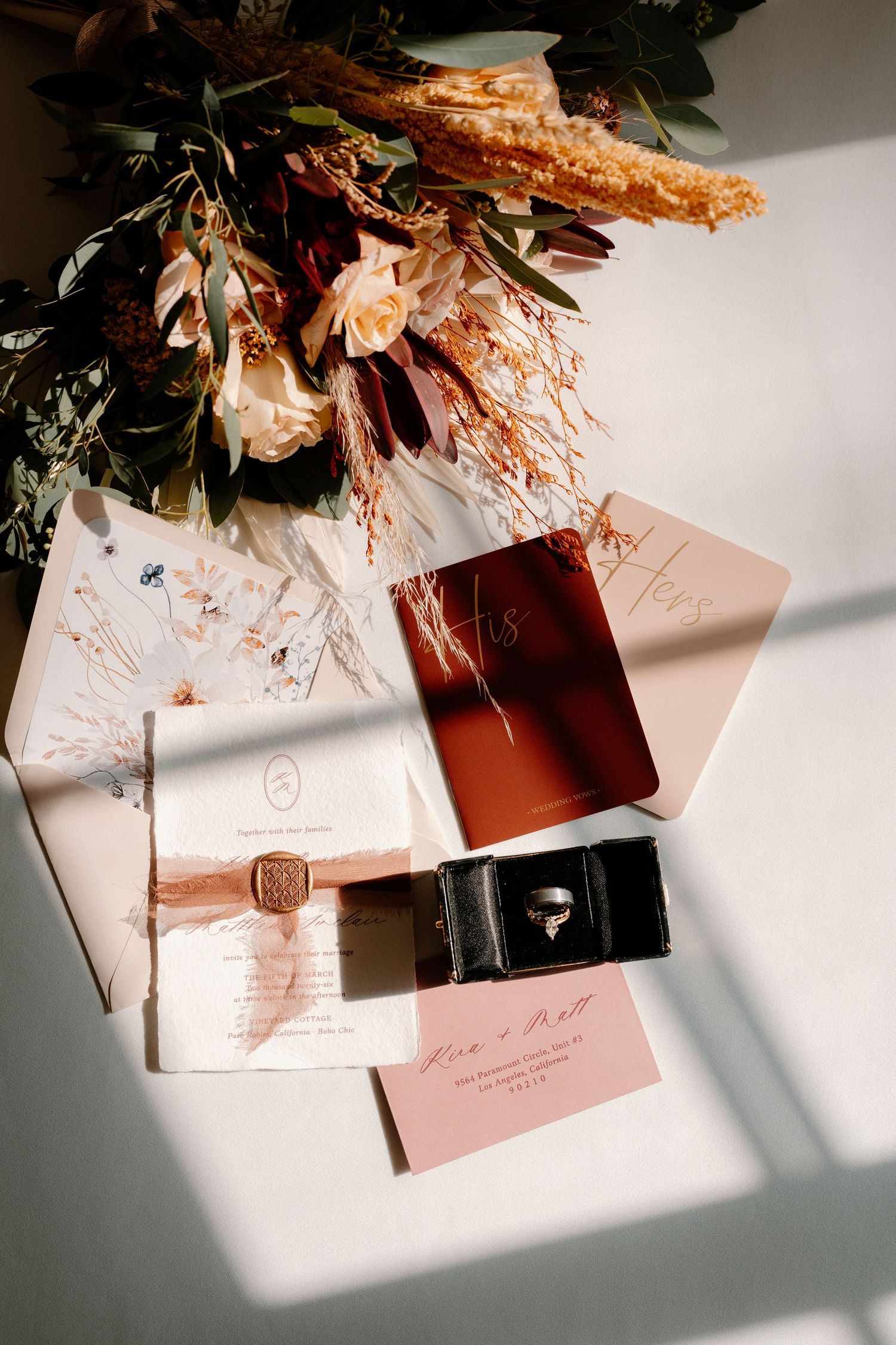 Flat lay of boho wedding stationery, ring box, and dried floral bouquet with warm terracotta tones in sunlight.