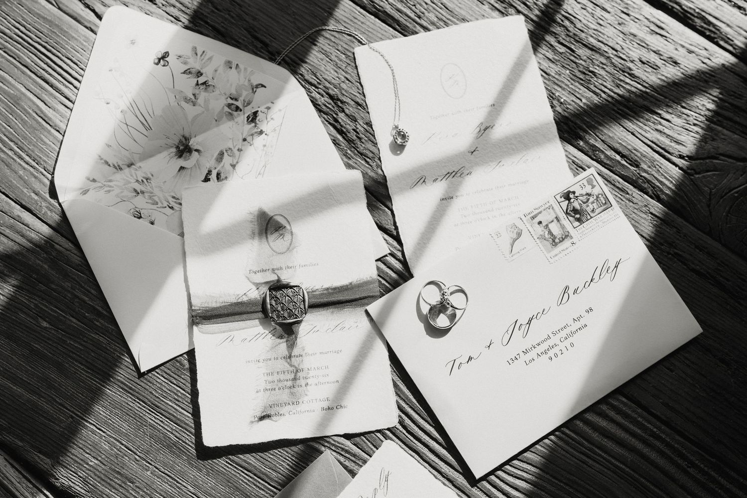 Black and white photo of elegant wedding invitation suite with wax seal, envelope, and rings on wooden surface.