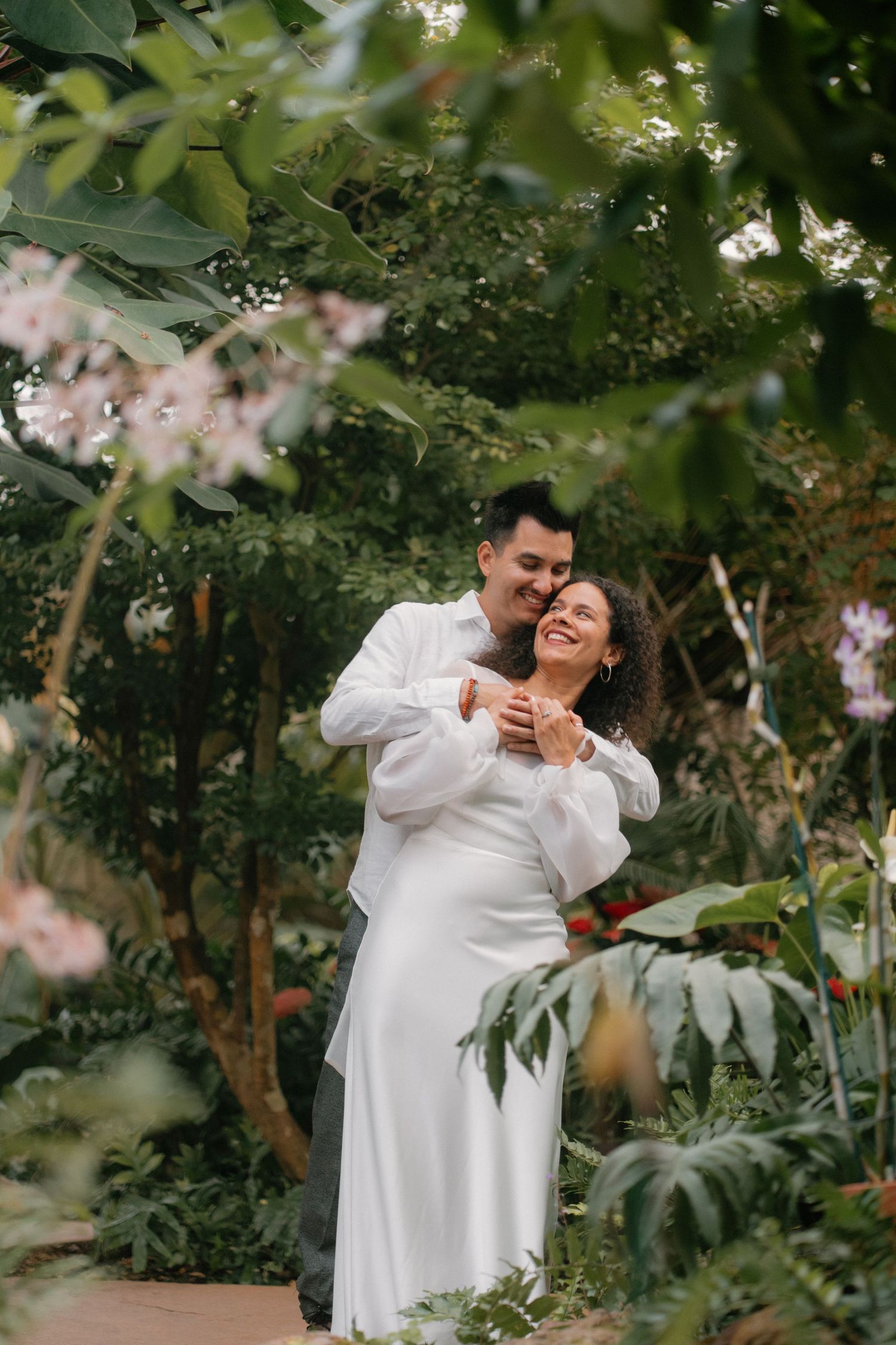 Couple embracing and smiling in a lush tropical garden, bride in white dress and groom in white shirt.
