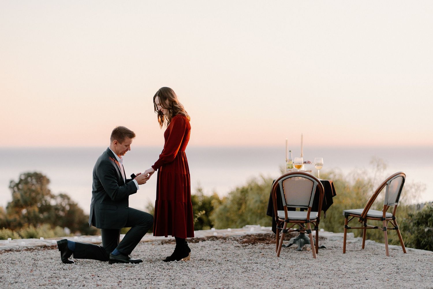 Romantic marriage proposal at sunset with man kneeling before woman in red dress.