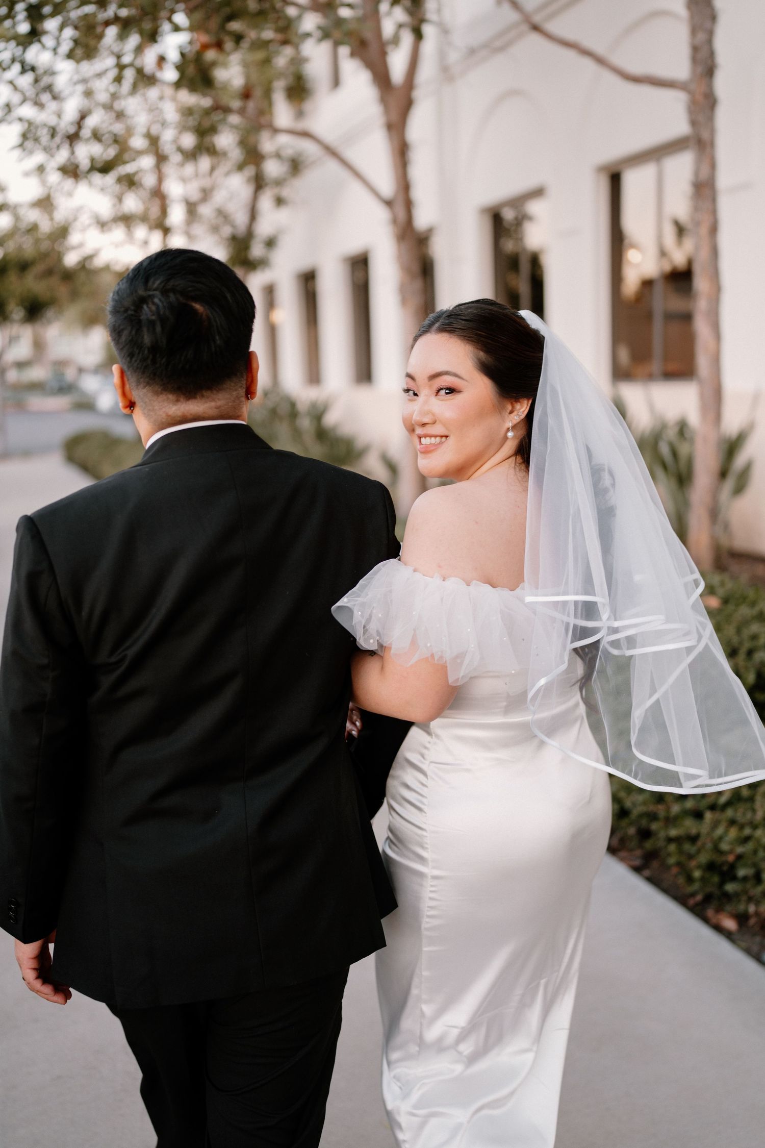 Smiling bride in white gown and veil walks arm-in-arm with groom outdoors.