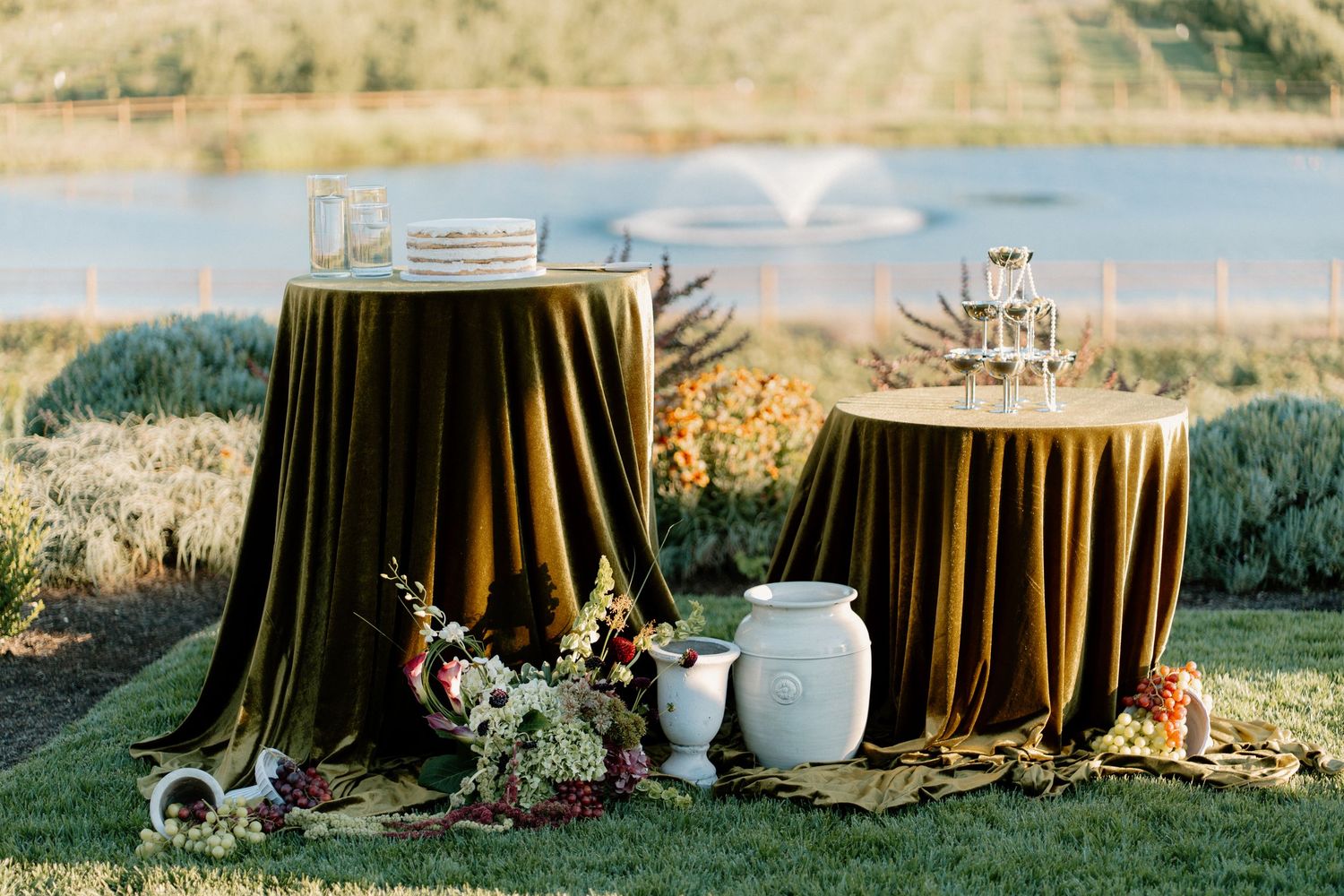 Elegant outdoor wedding reception tables draped in olive velvet with floral arrangements and glassware by a lake.