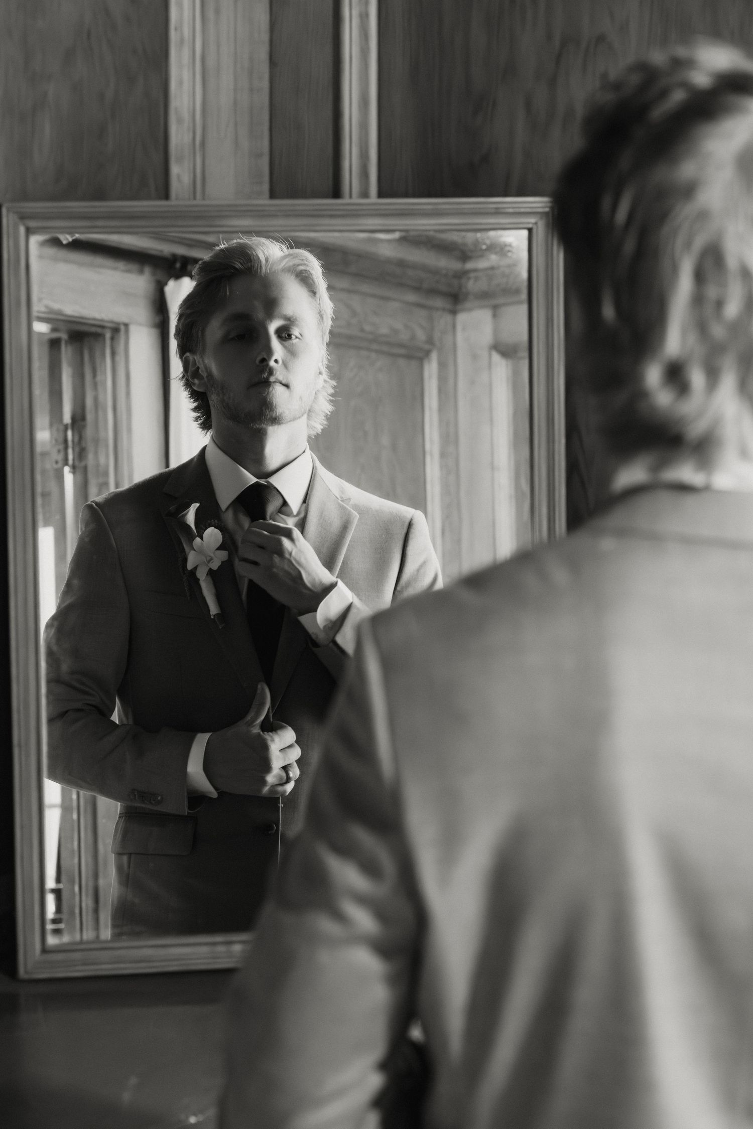 Black and white photo of a groom adjusting his tie in a mirror while wearing a suit with boutonniere.