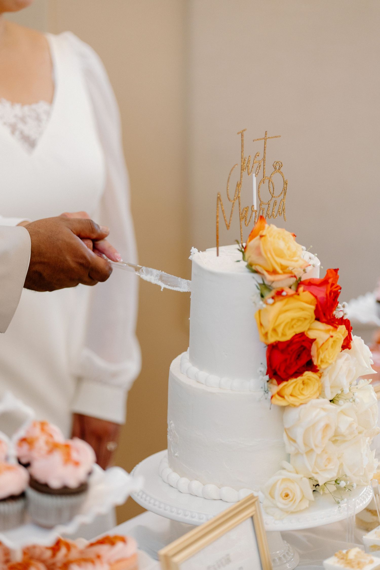 Bride cutting elegant three-tier white wedding cake decorated with colorful roses and Just Married topper.