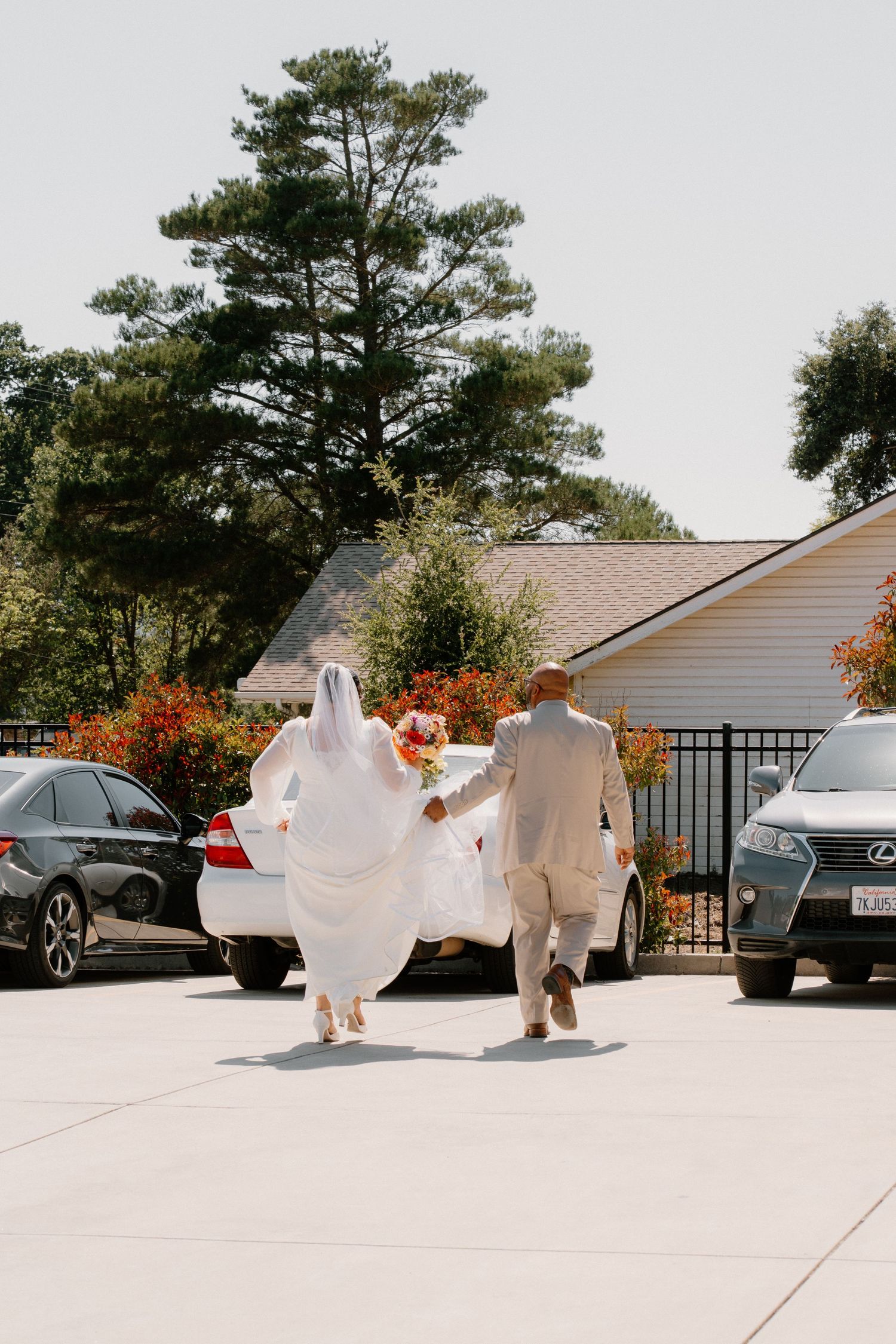 Bride in white gown and veil walking with groom in tan suit through sunny outdoor parking area.