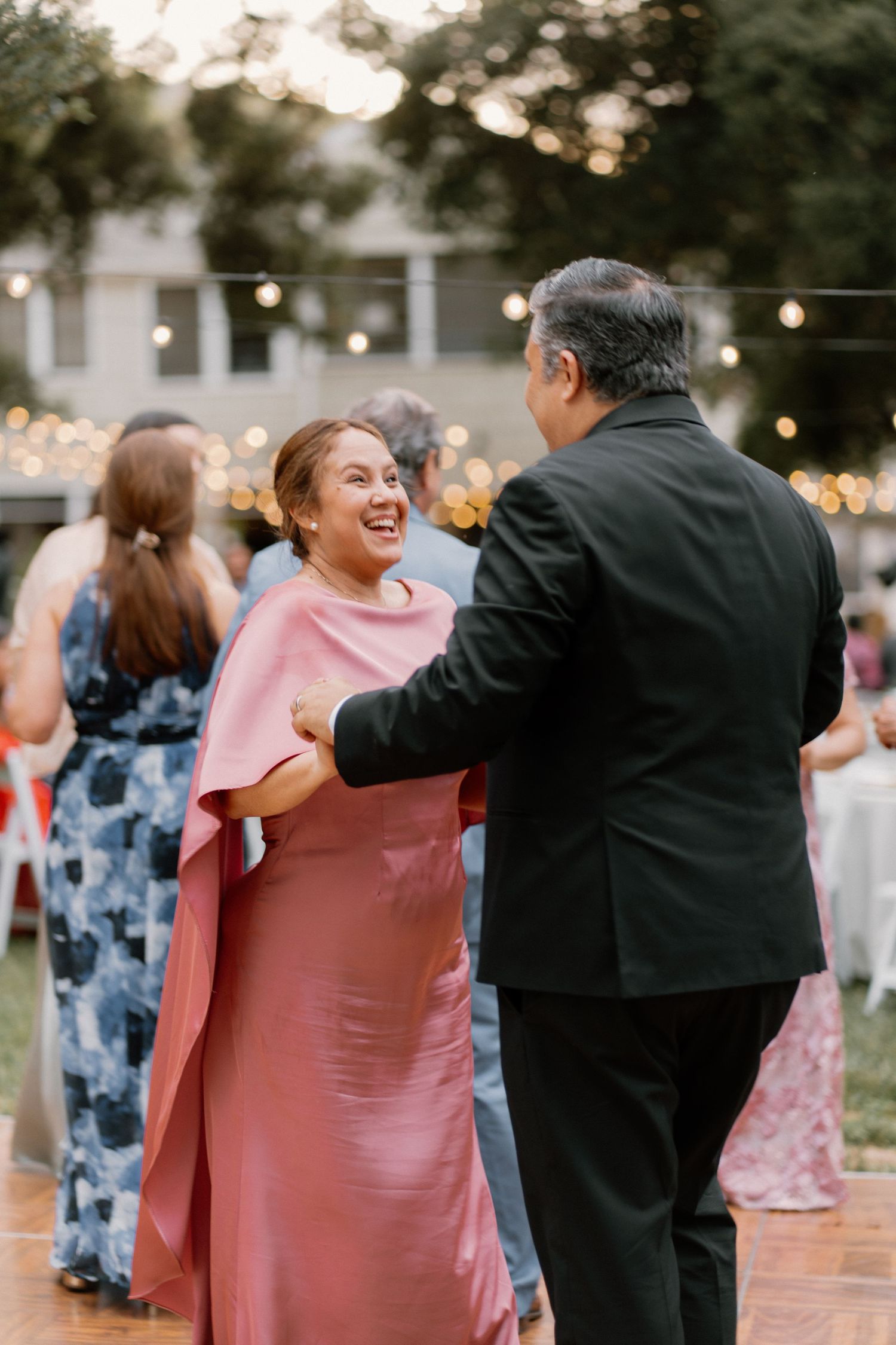 A joyful woman in a pink gown dances with a man in a black suit at an outdoor wedding reception.
