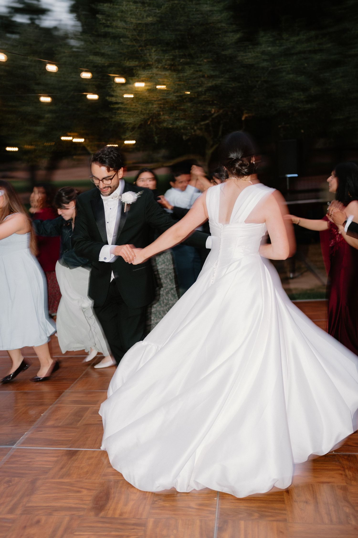 Bride in flowing white ball gown twirls with groom in black tuxedo on outdoor wedding dance floor.