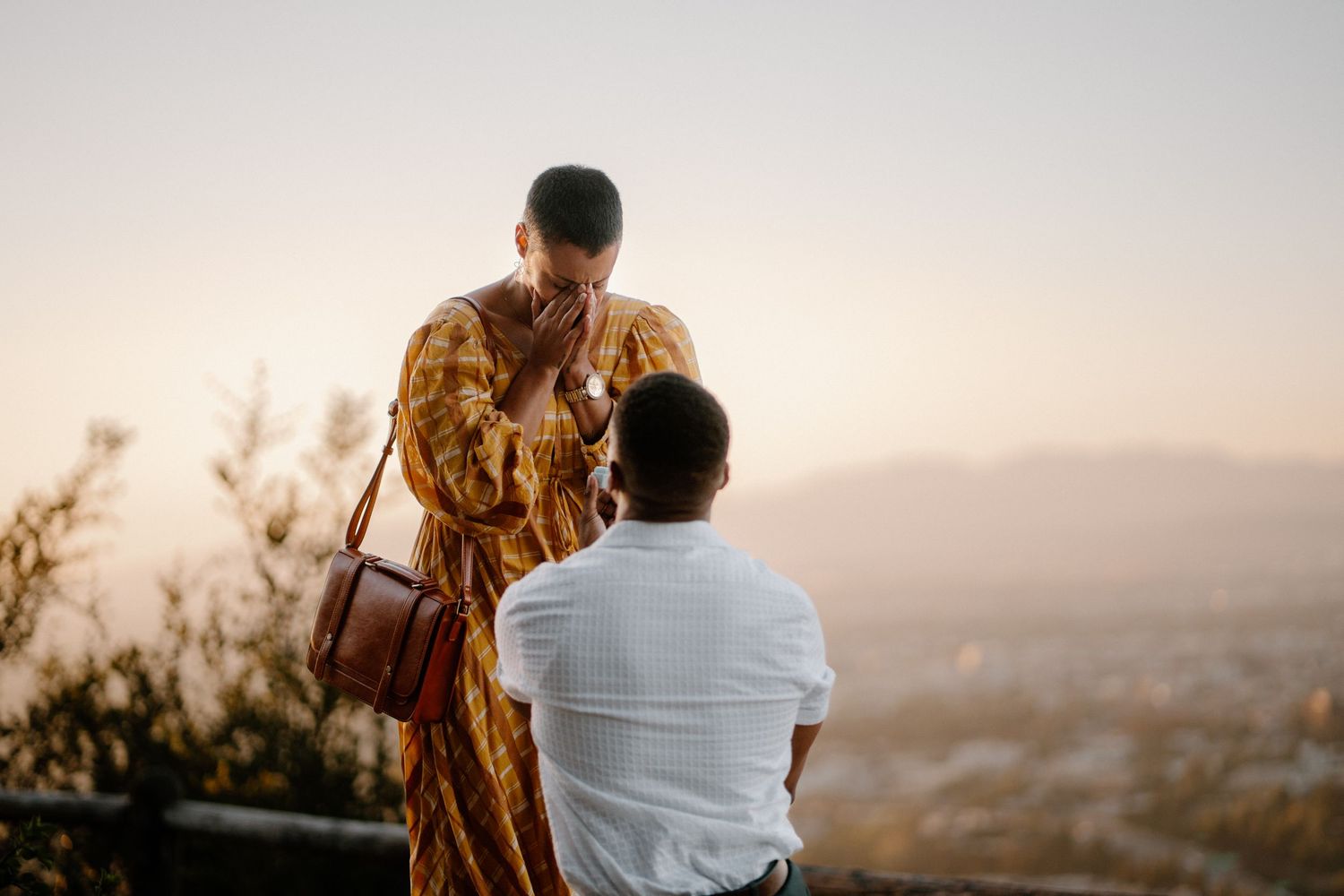 Woman in yellow dress holding hands with man at a scenic overlook during golden hour sunset.
