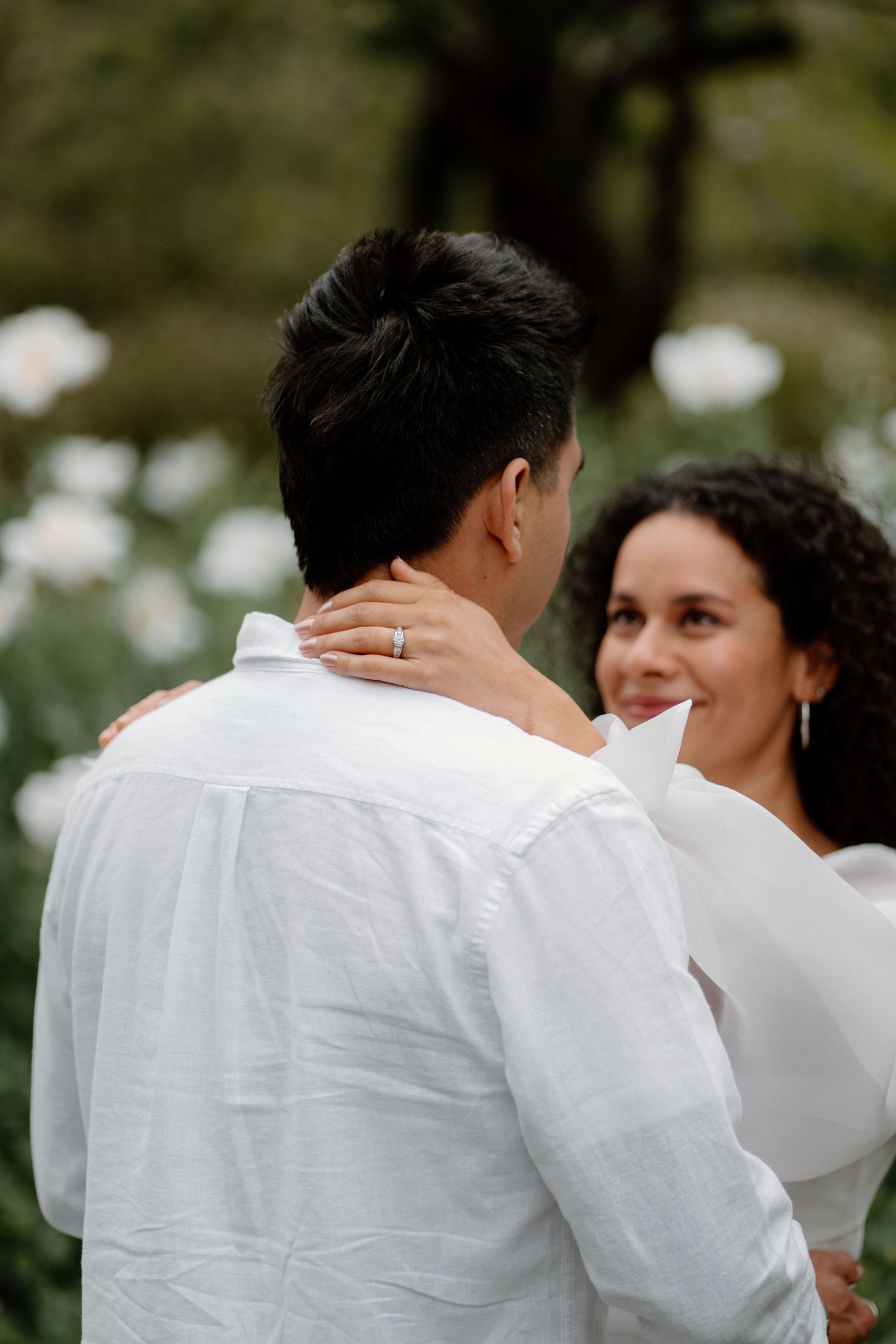 Woman smiling as she embraces her fiance from behind near white flowers during engagement shoot.
