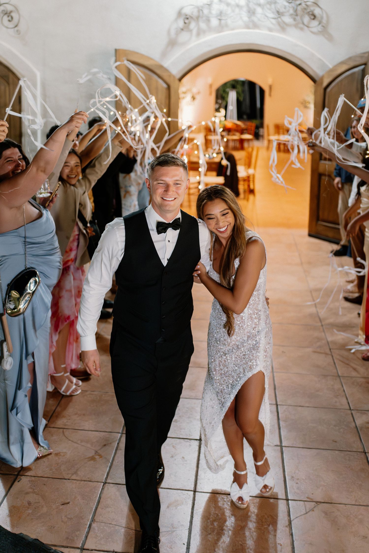 Bride and groom walk through ribbon send-off tunnel surrounded by cheering wedding guests in elegant venue.