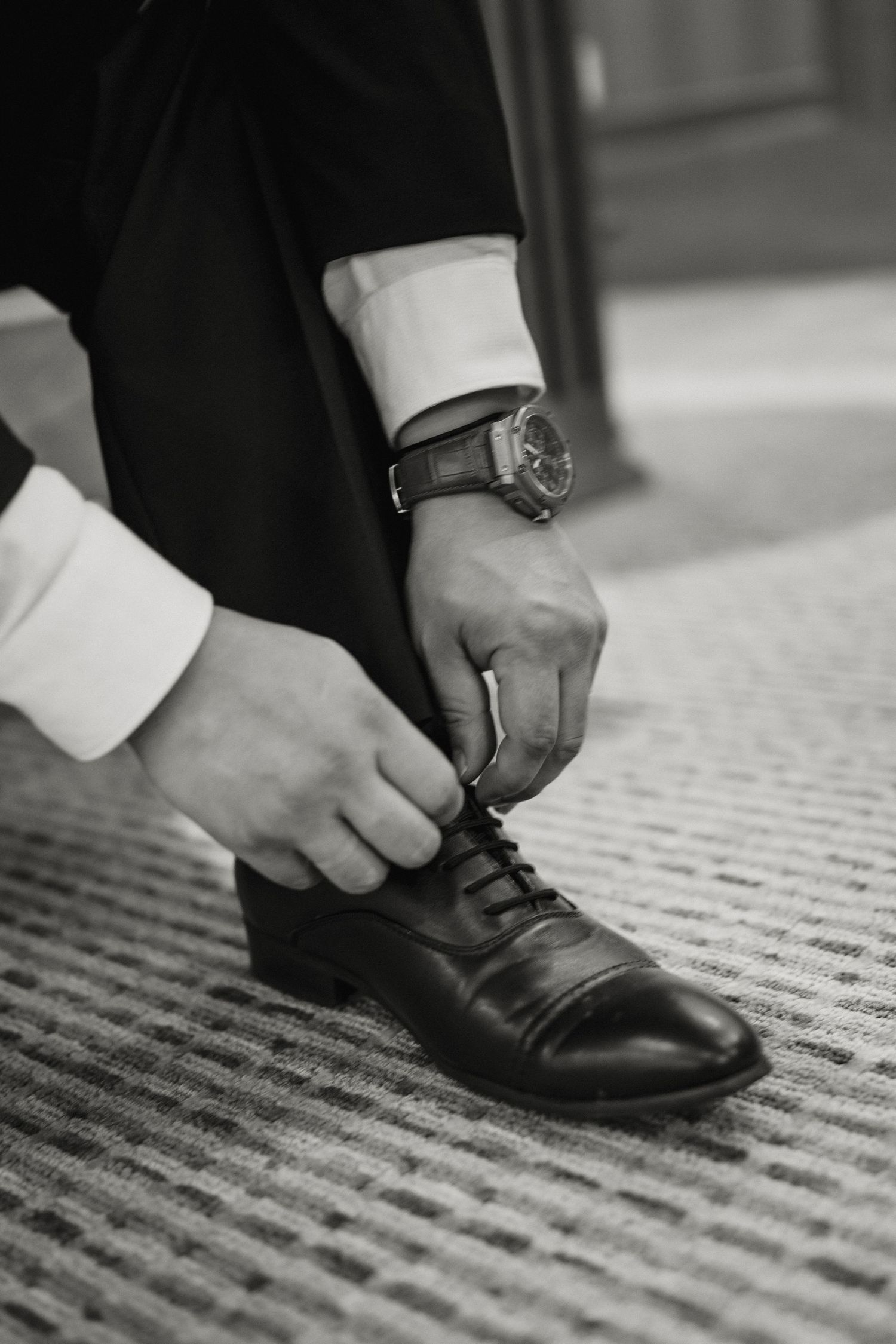 Black and white close-up of groom tying his dress shoes while wearing a watch on his wrist.