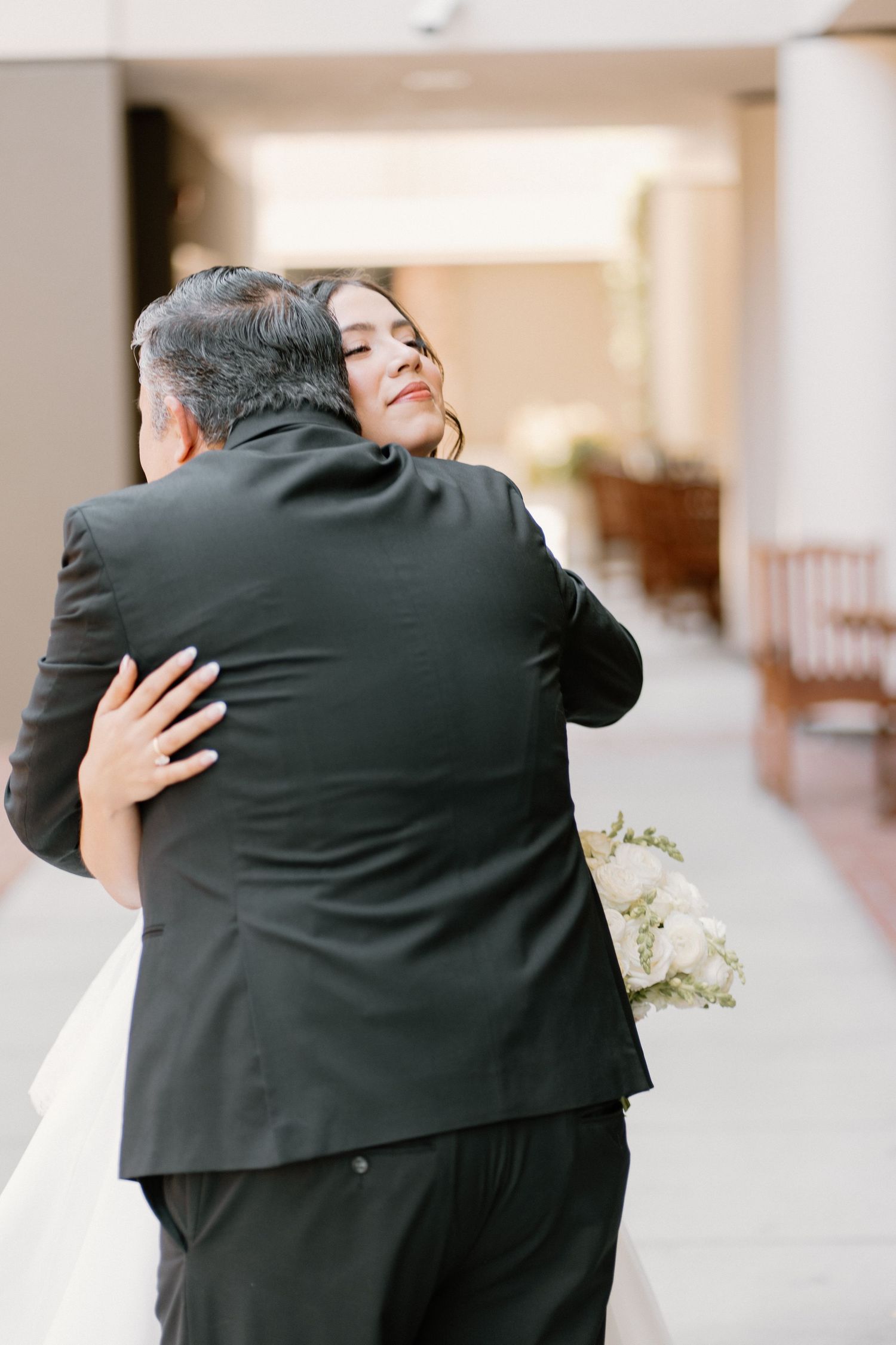 Emotional bride in white gown embracing a man in a black suit while holding a white bouquet.