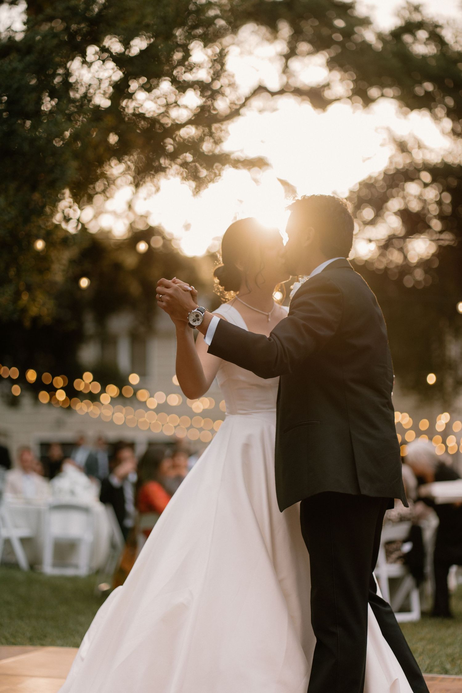 Bride and groom share first dance outdoors at golden hour, backlit by sunset with string lights glowing in trees.