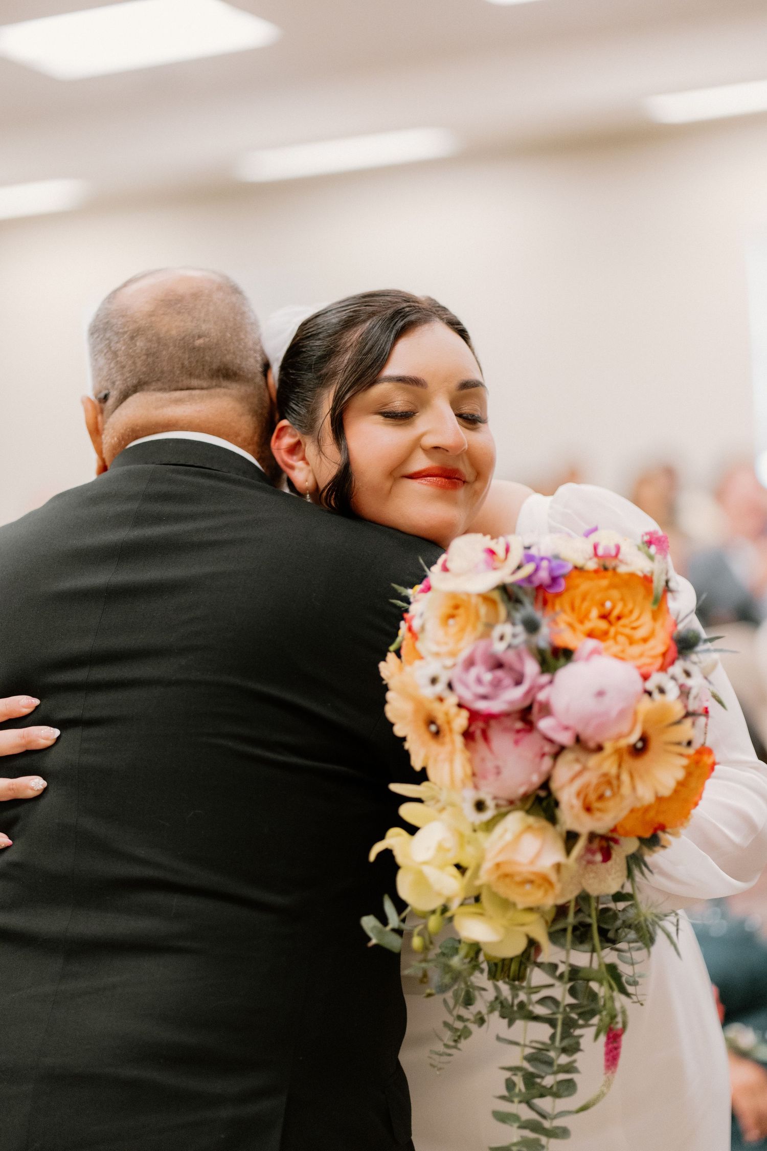 Bride in white gown hugging guest while holding vibrant orange and pink wedding bouquet at ceremony.