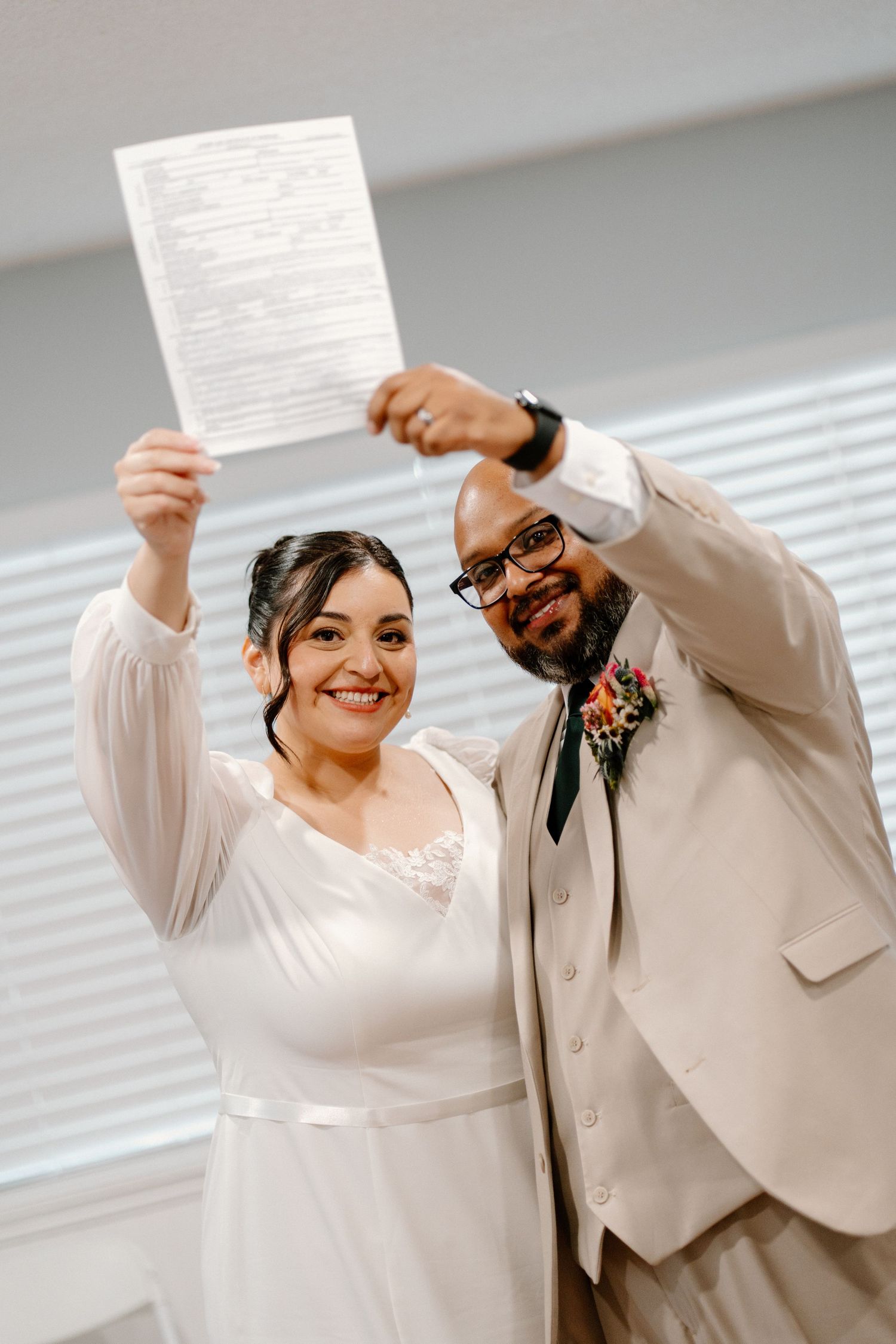 Joyful newlyweds in wedding attire proudly hold up their signed marriage certificate together.