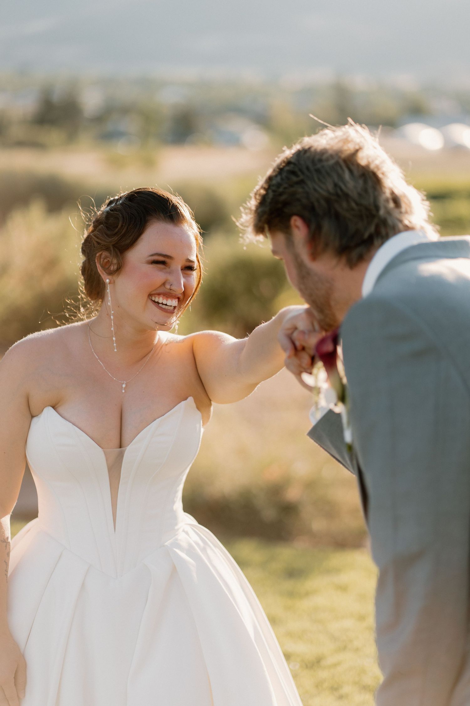 Joyful bride in strapless ballgown laughing as groom kisses her hand at golden hour.