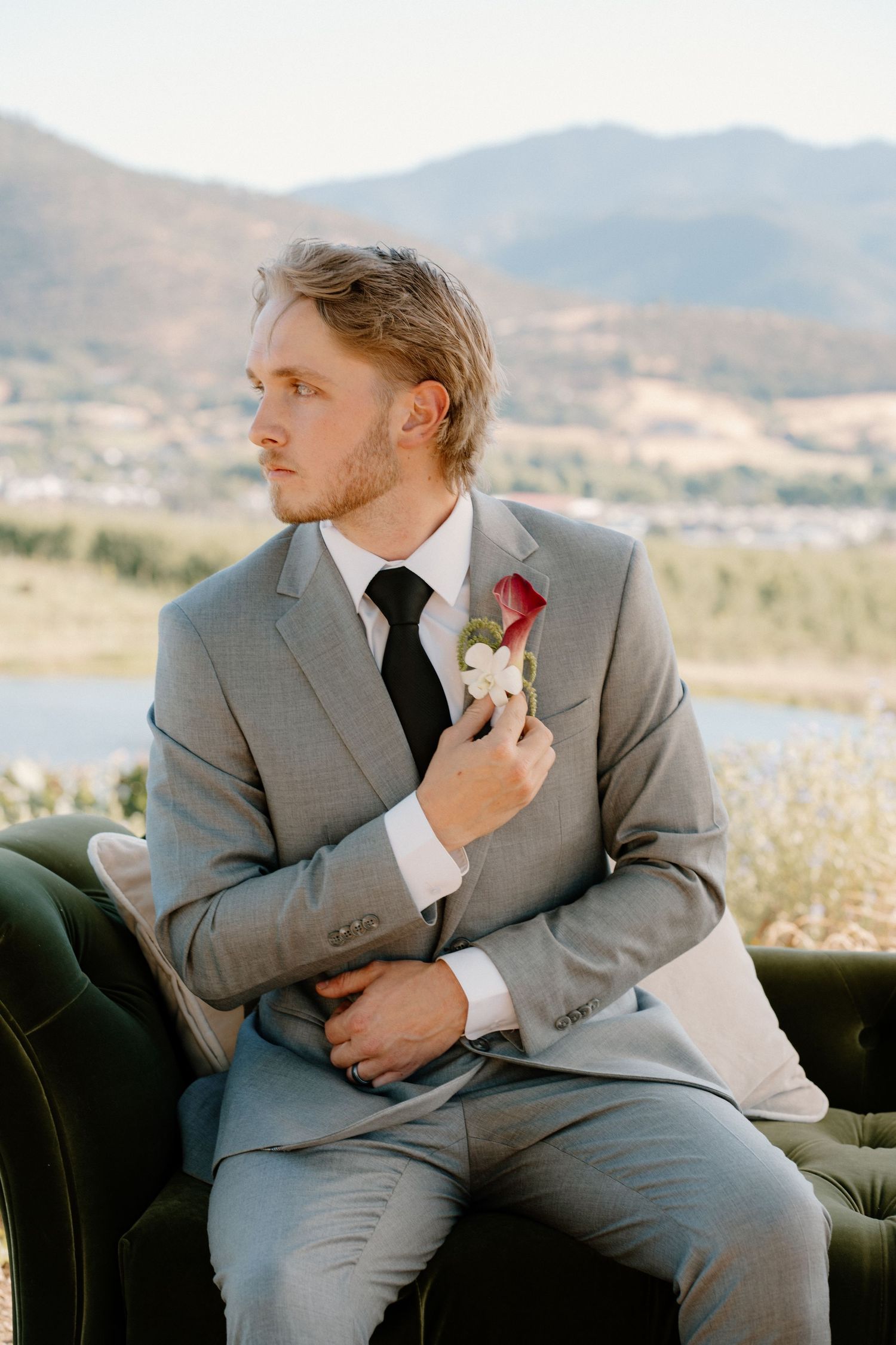 Groom in gray suit with boutonniere sits on green velvet chair with mountain landscape backdrop.