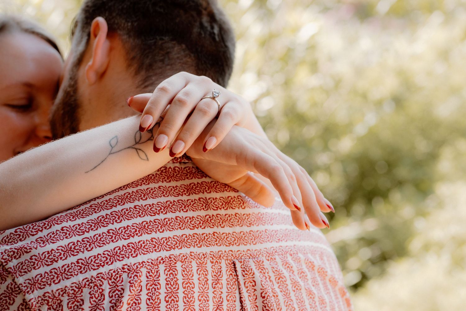 Couple sharing a tender embrace outdoors, woman with tattoo and rings wrapping arms around man in striped shirt.