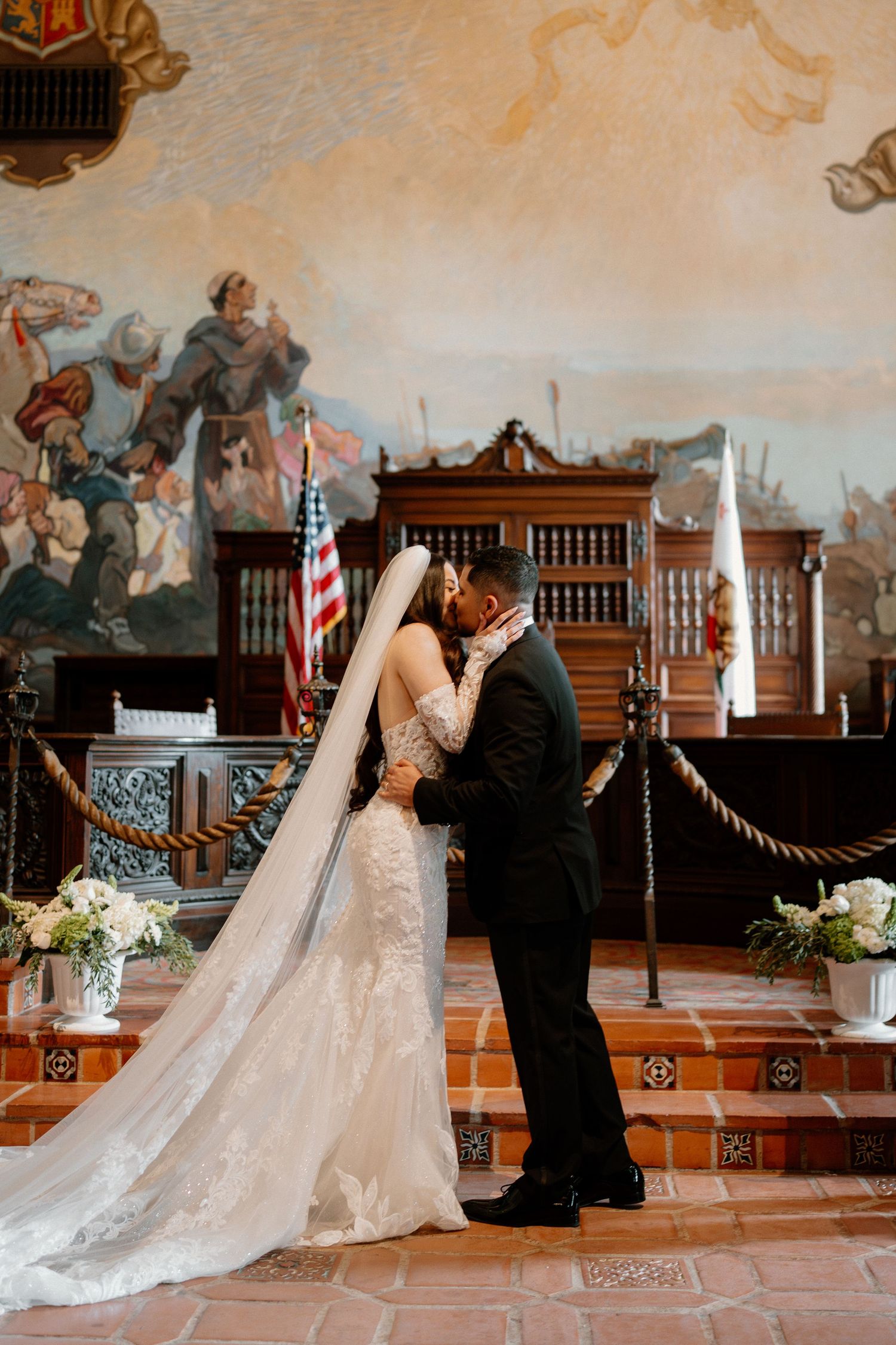 Bride and groom share first kiss at altar with large historical mural and American flag backdrop.