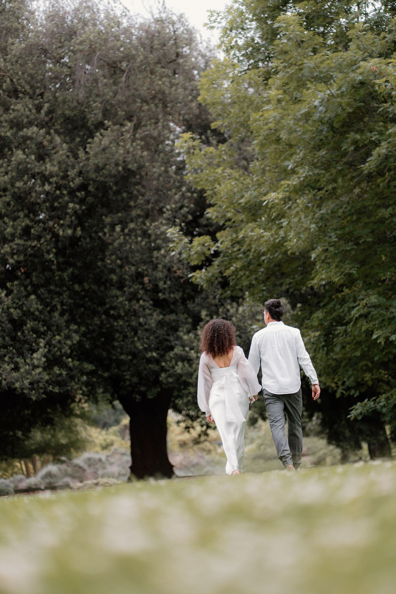 Couple walking hand in hand away from camera through a scenic tree-lined park path.