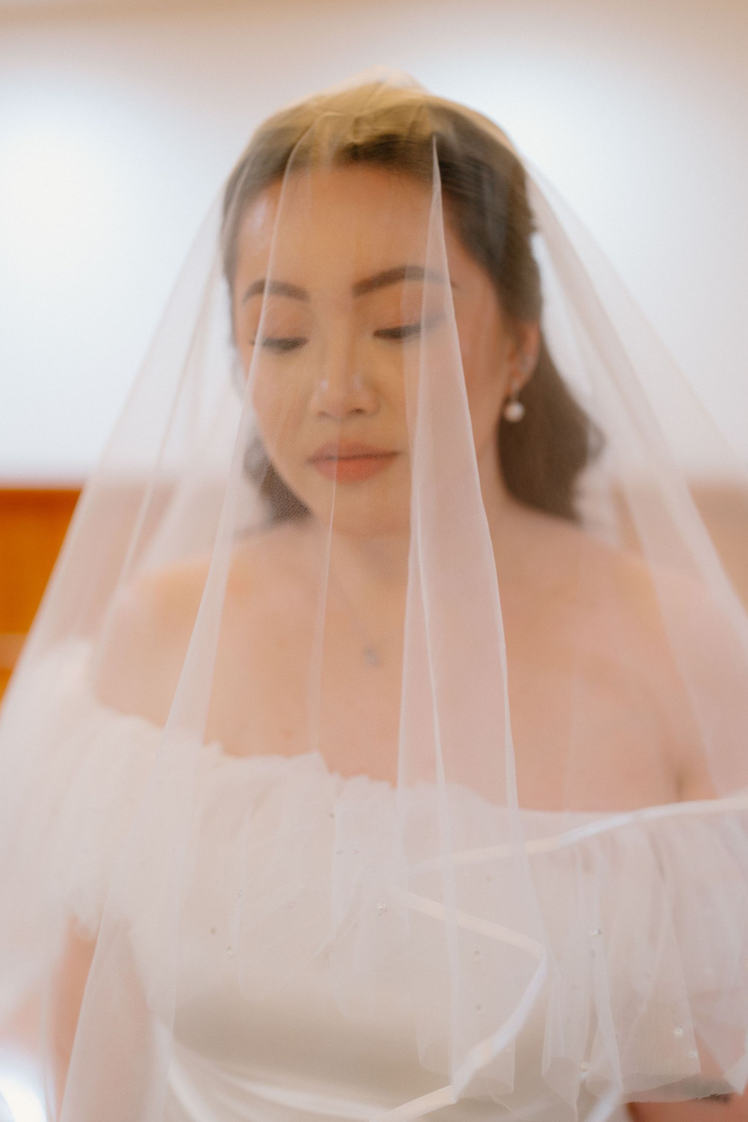 Elegant bride with eyes closed, draped in a sheer white veil, in a softly lit indoor setting.