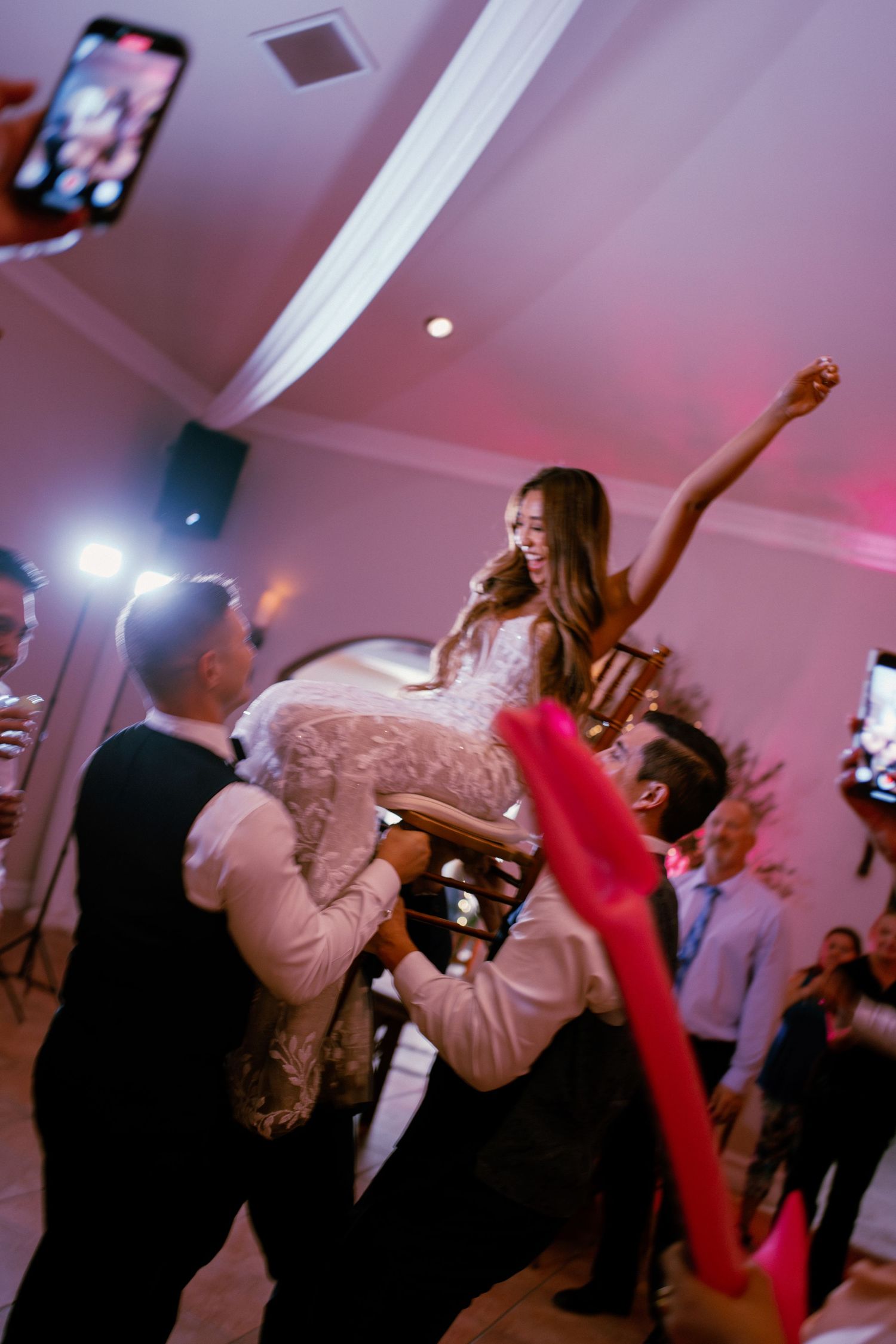 Joyful bride being lifted by groomsmen on a chair during a lively wedding reception dance celebration.