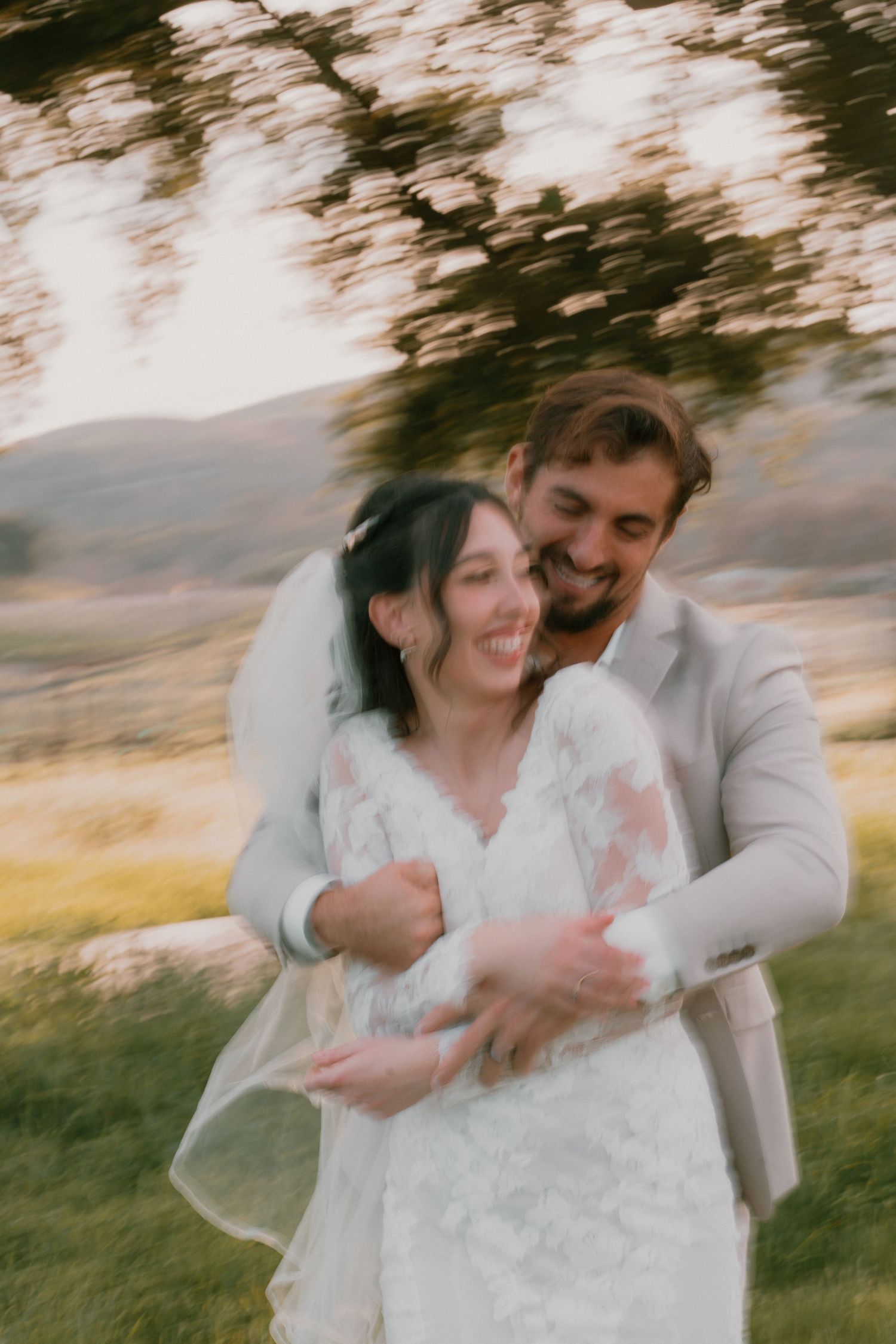 Artistic motion-blur wedding photo of laughing couple embracing outdoors with lush green landscape behind them.